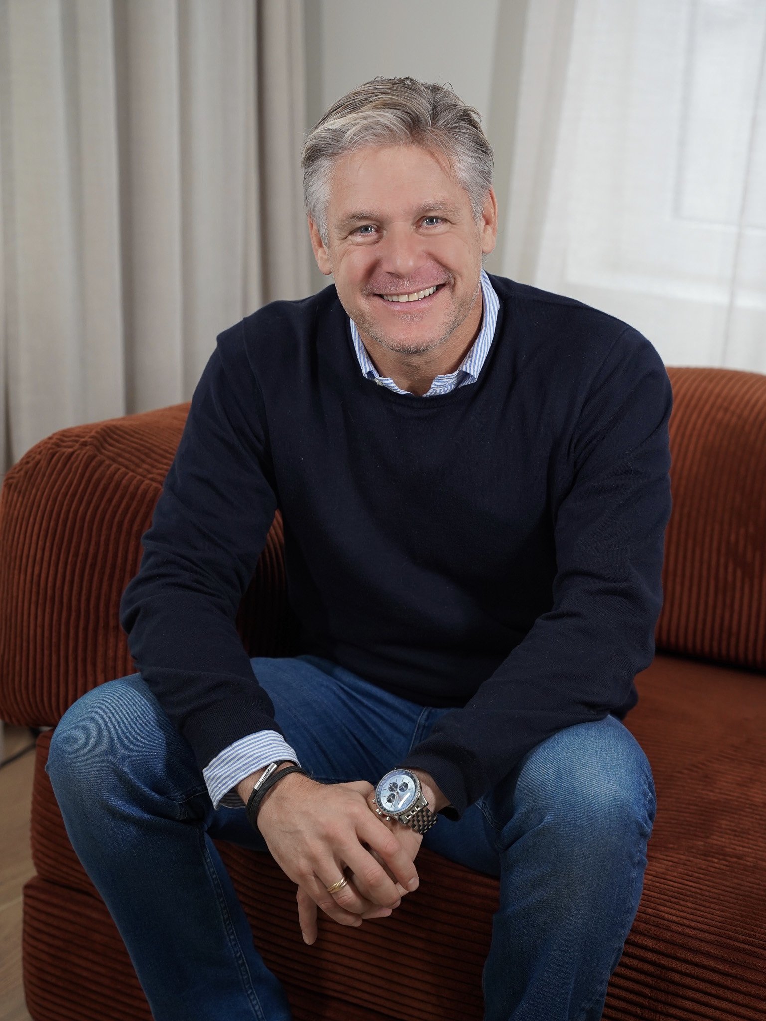 Smiling man with gray hair wearing a gray sweater and white shirt, standing indoors with a blurred background.