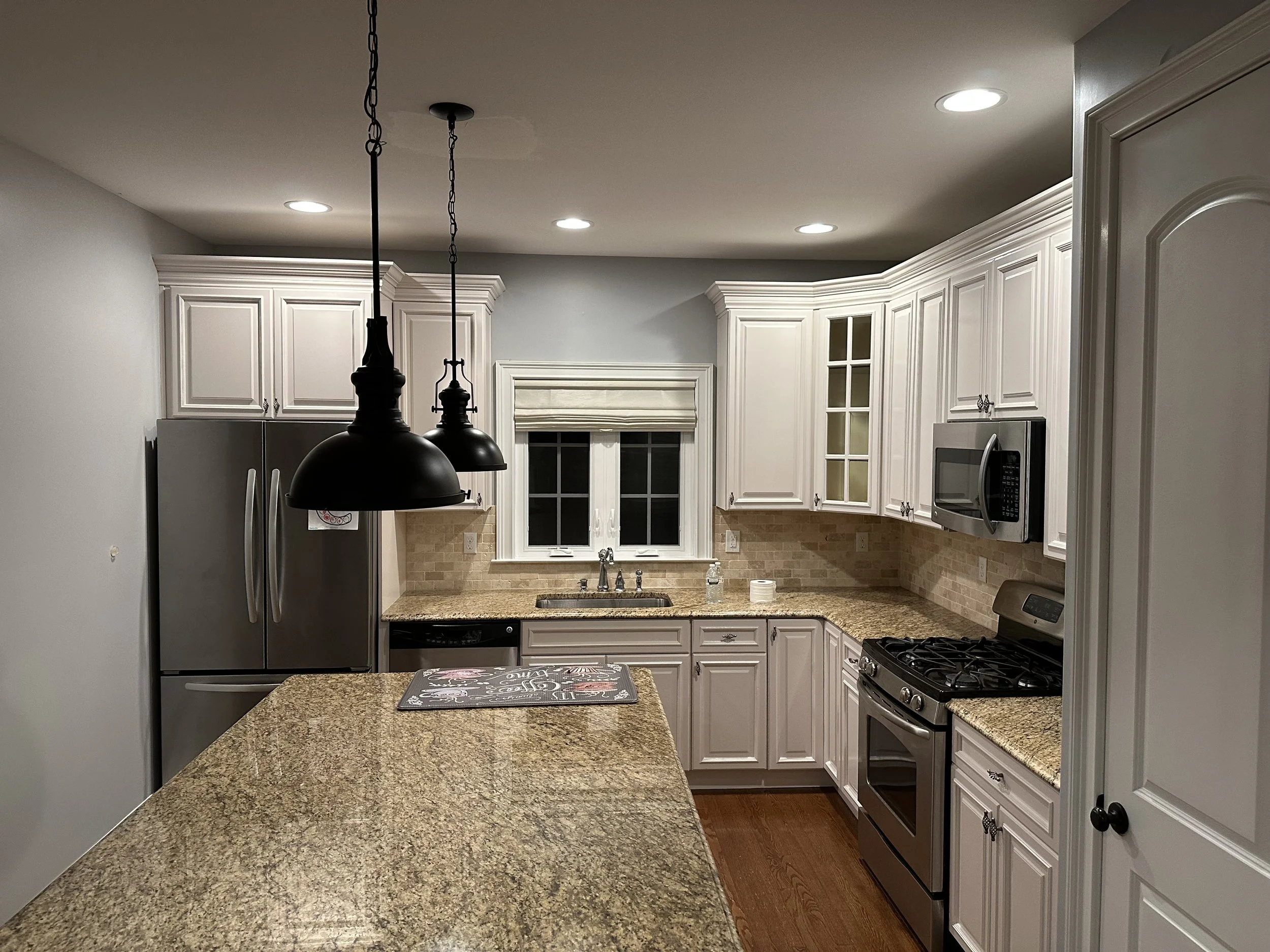 Kitchen with white cabinets, granite countertops, stainless steel appliances, and a window above the sink. Two black pendant lights hang above a kitchen island.