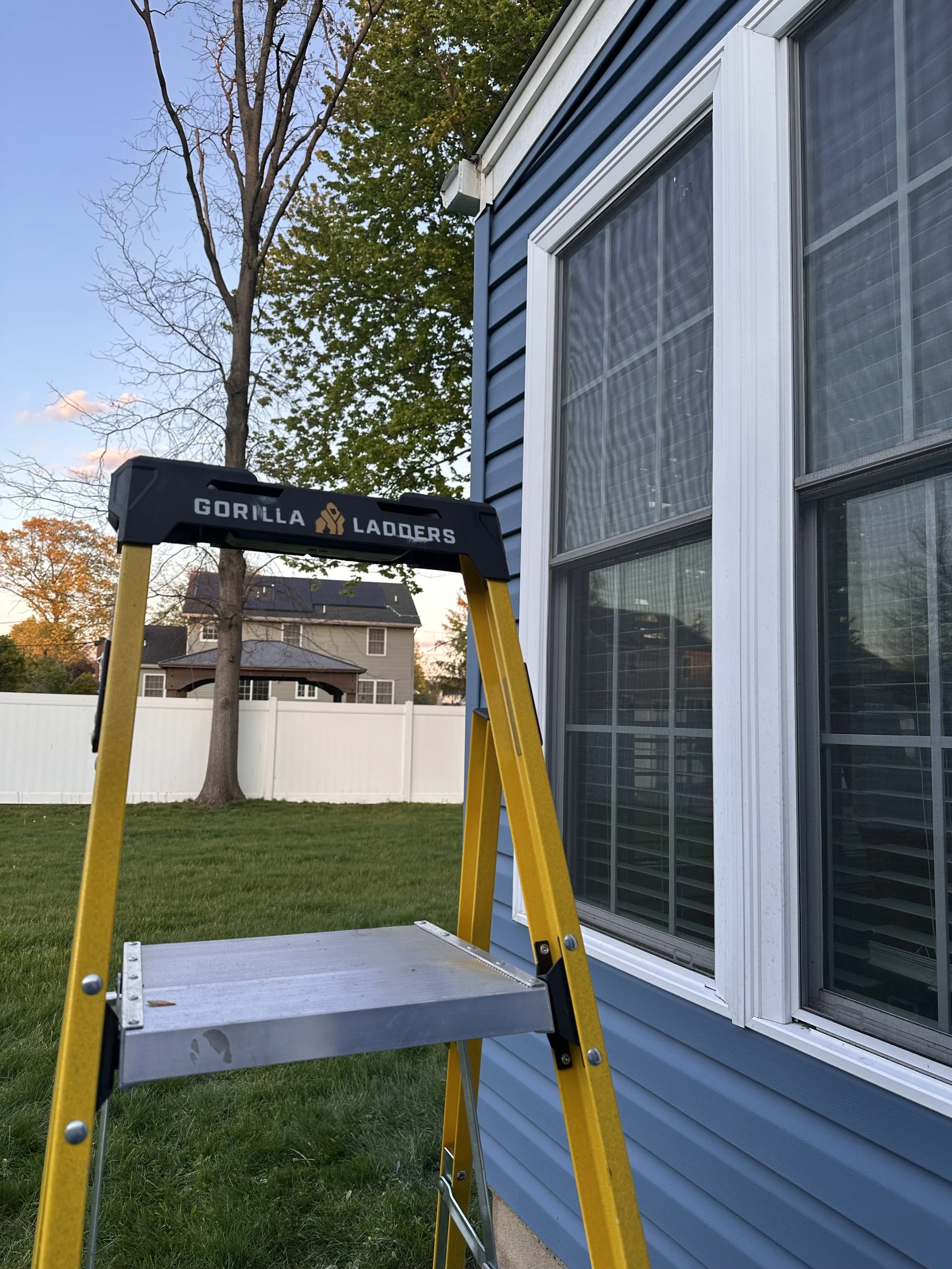 A yellow Gorilla Ladders step ladder set up outside a house with light blue siding and large windows, in a backyard with green grass, trees, and a white fence.