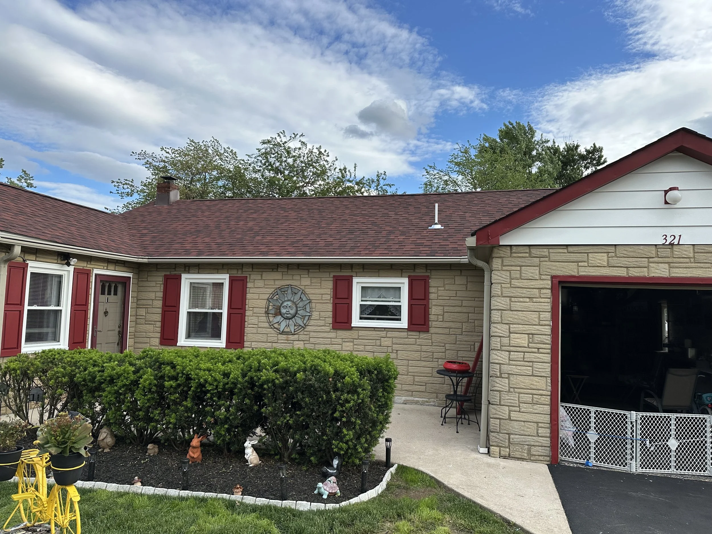 A house with a red shingle roof, stone siding, red window shutters, and a sun decoration on the front wall, with a garden in front containing bushes, figurines, and plants, and a bright blue sky with clouds overhead.