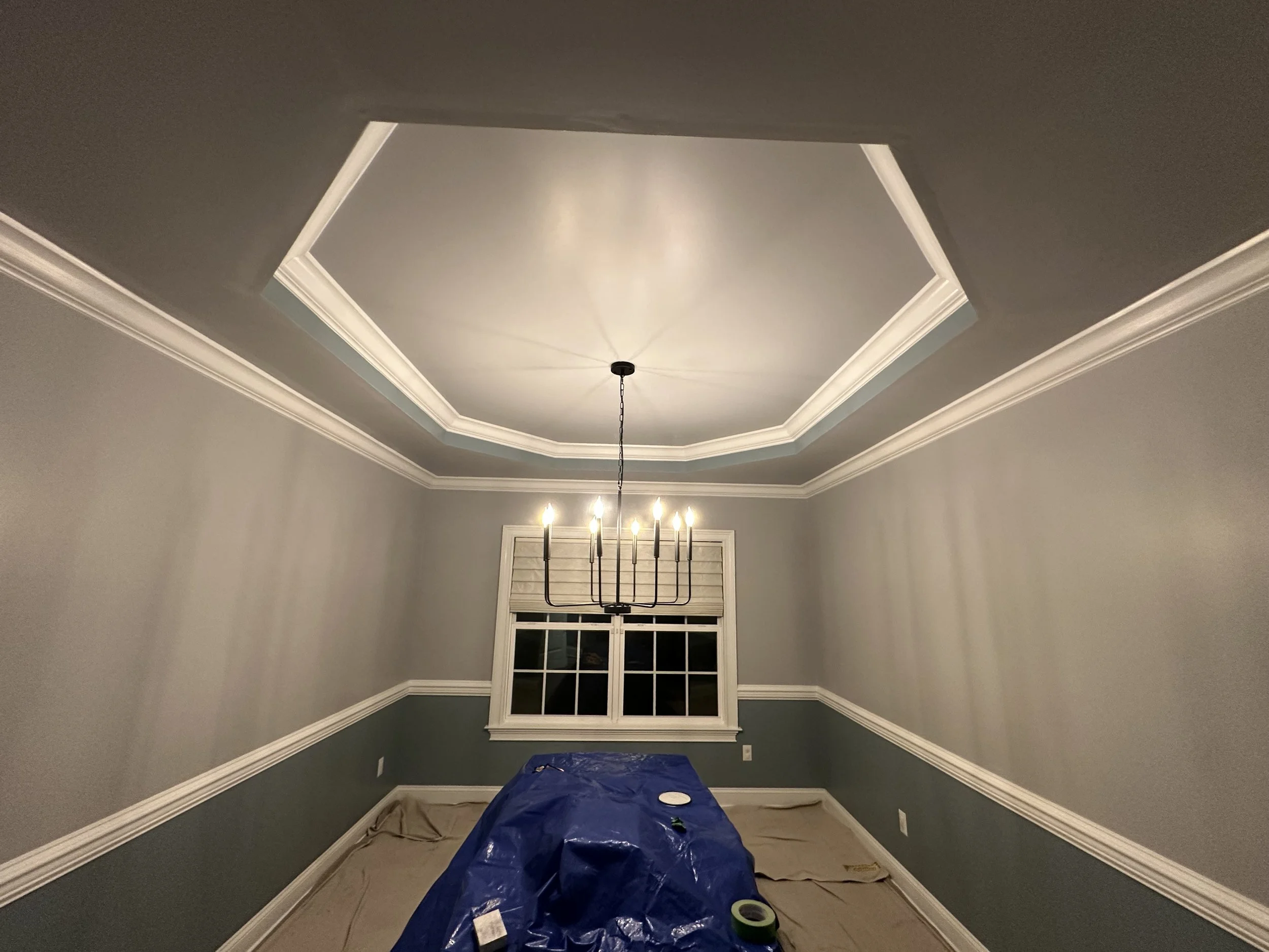 A room with tray ceiling and crown molding, chandelier hanging from chain, window with blinds, covered with painter's tarp and painter's tape on the floor, suggesting ongoing painting work.