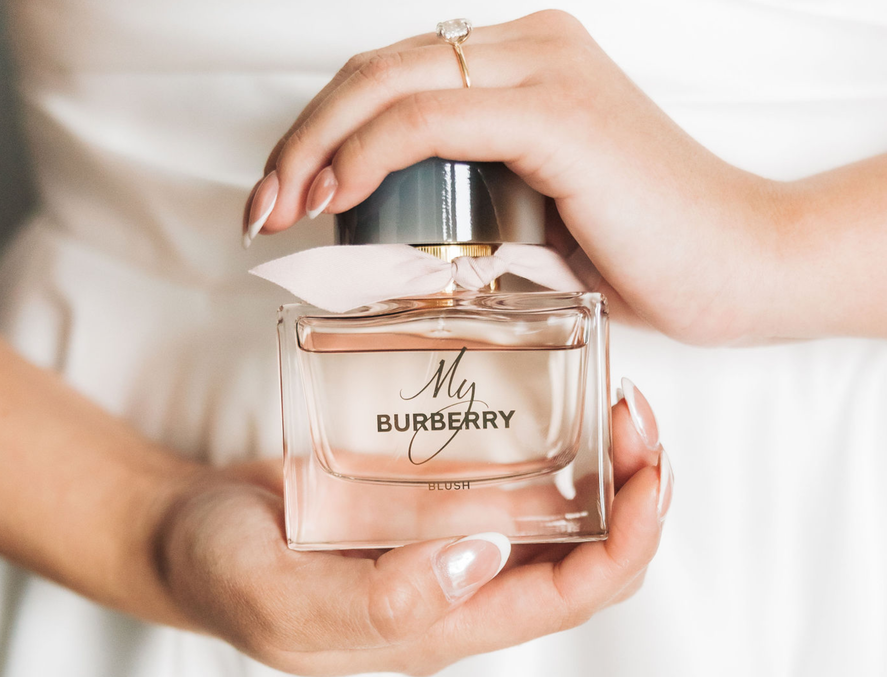 A hand holding a bottle of Burberry perfume labeled "My Burberry" with a white ribbon around the neck of the bottle.