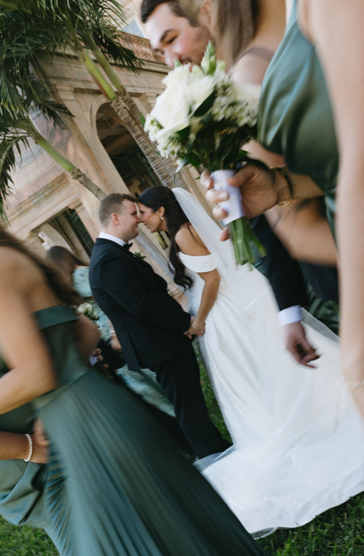 Bride and groom holding hands and smiling at each other during their wedding ceremony outdoors, surrounded by wedding guests.