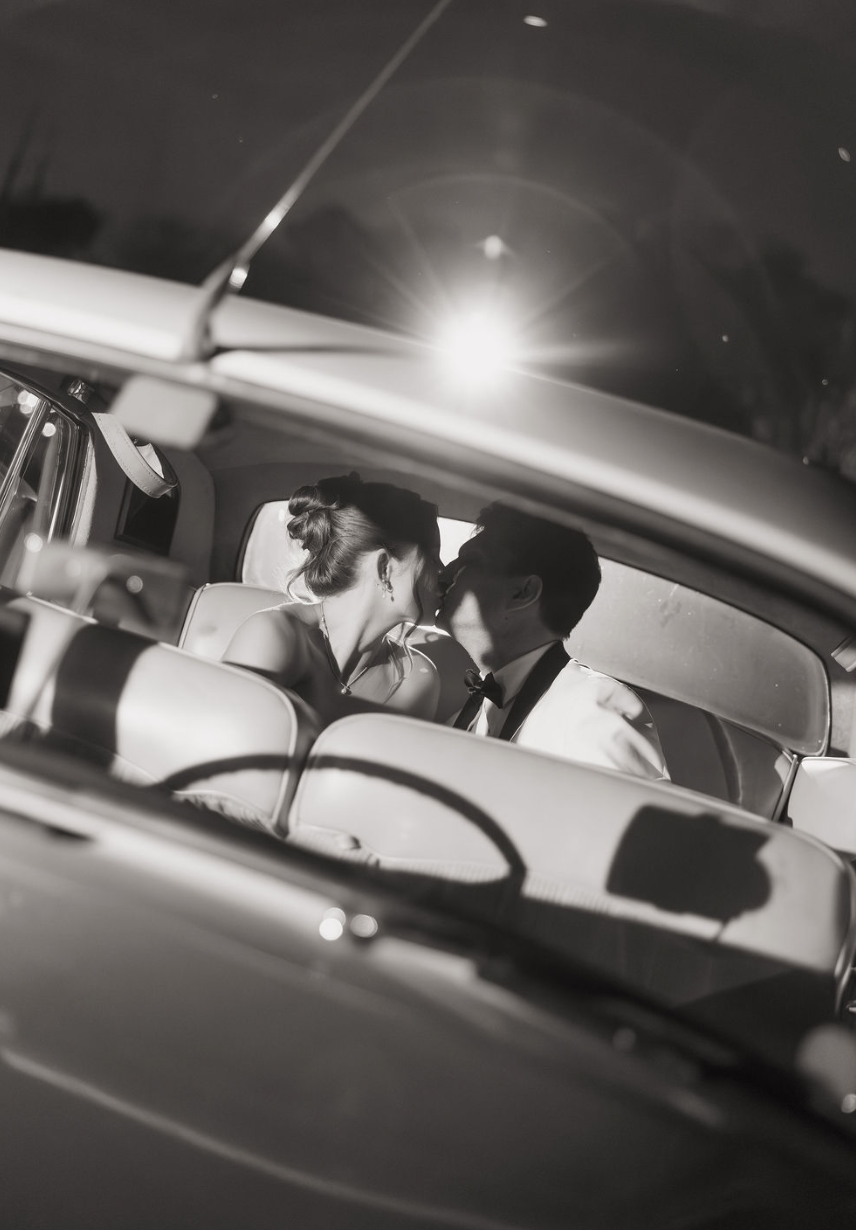 A black and white photo of a couple in formal attire sharing a kiss inside a vintage car.