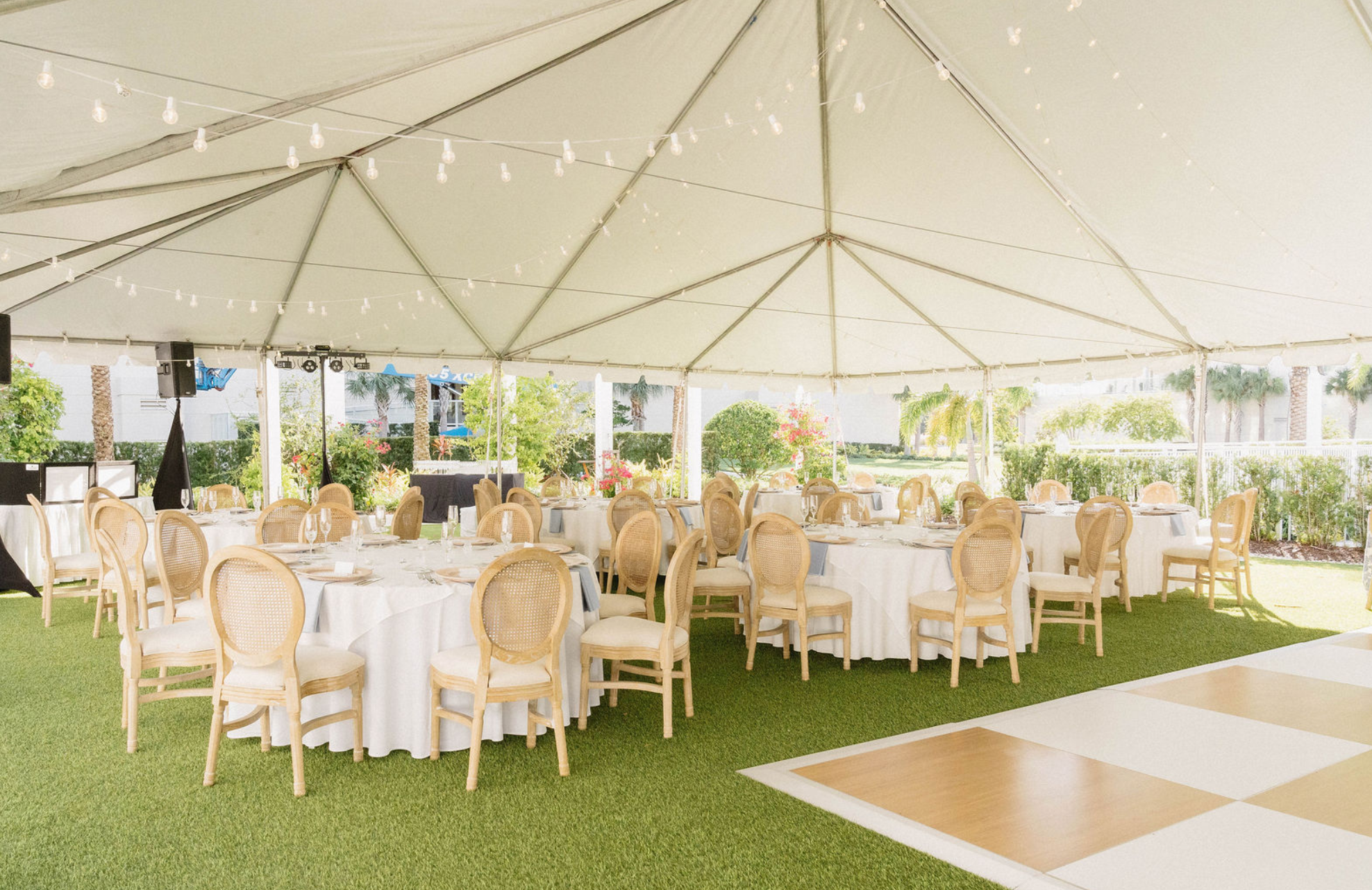 Outdoor event tent with round tables and chairs, decorated with string lights and surrounded by greenery.