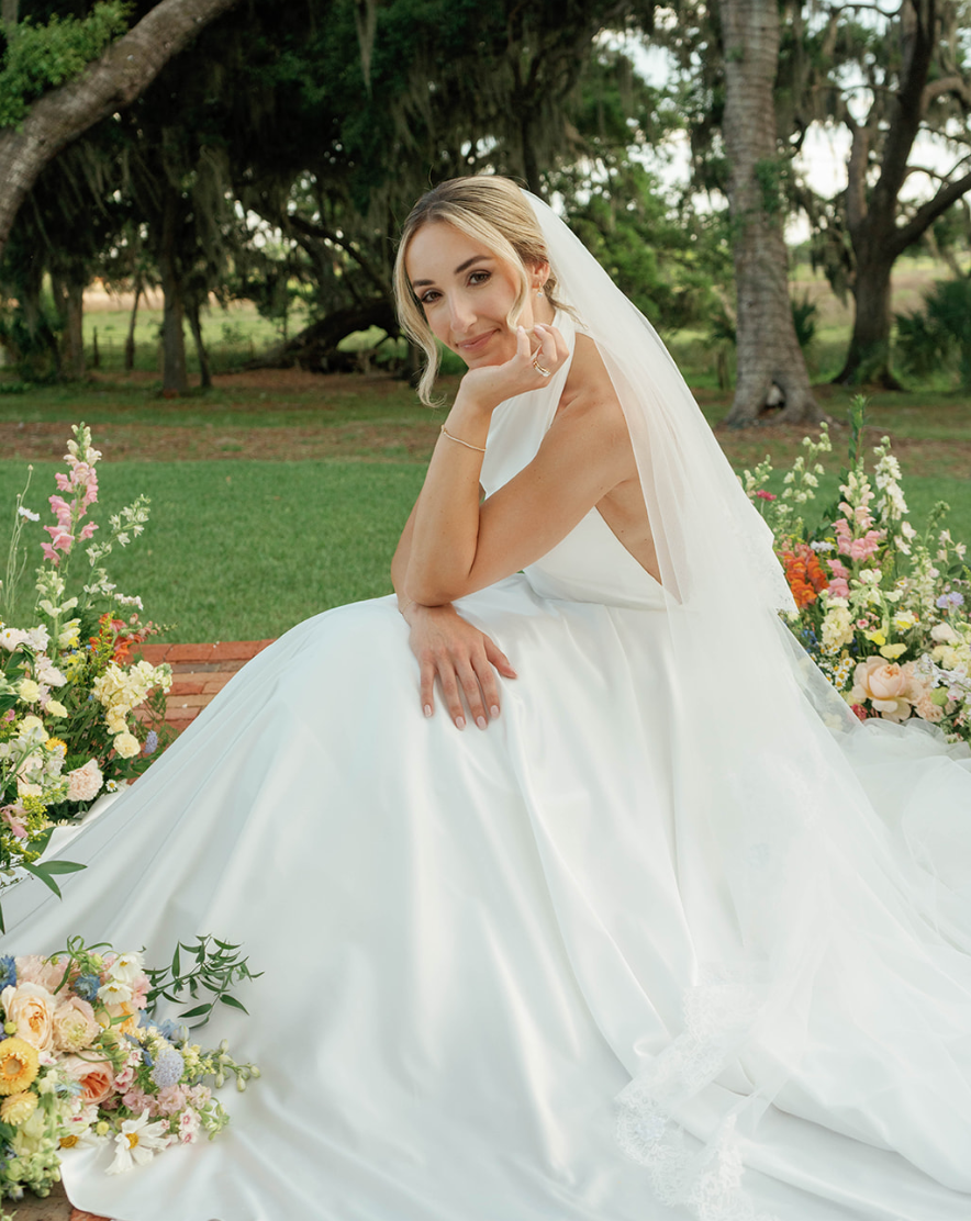 Brunette bride in white wedding gown sitting outside among flowers with trees in the background.