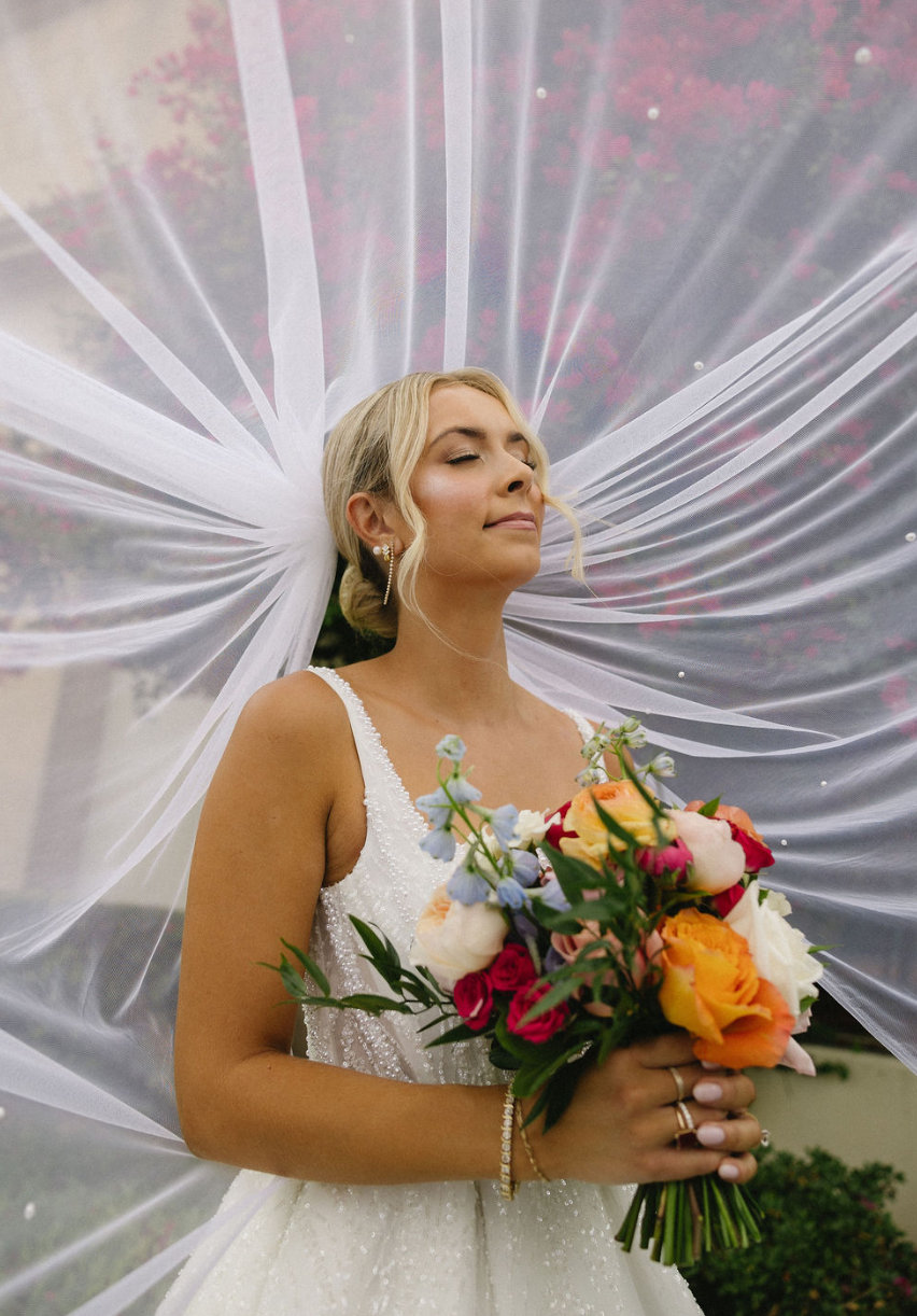 Bride with closed eyes holding a colorful bouquet of flowers, wearing a white wedding dress and veil outdoors.