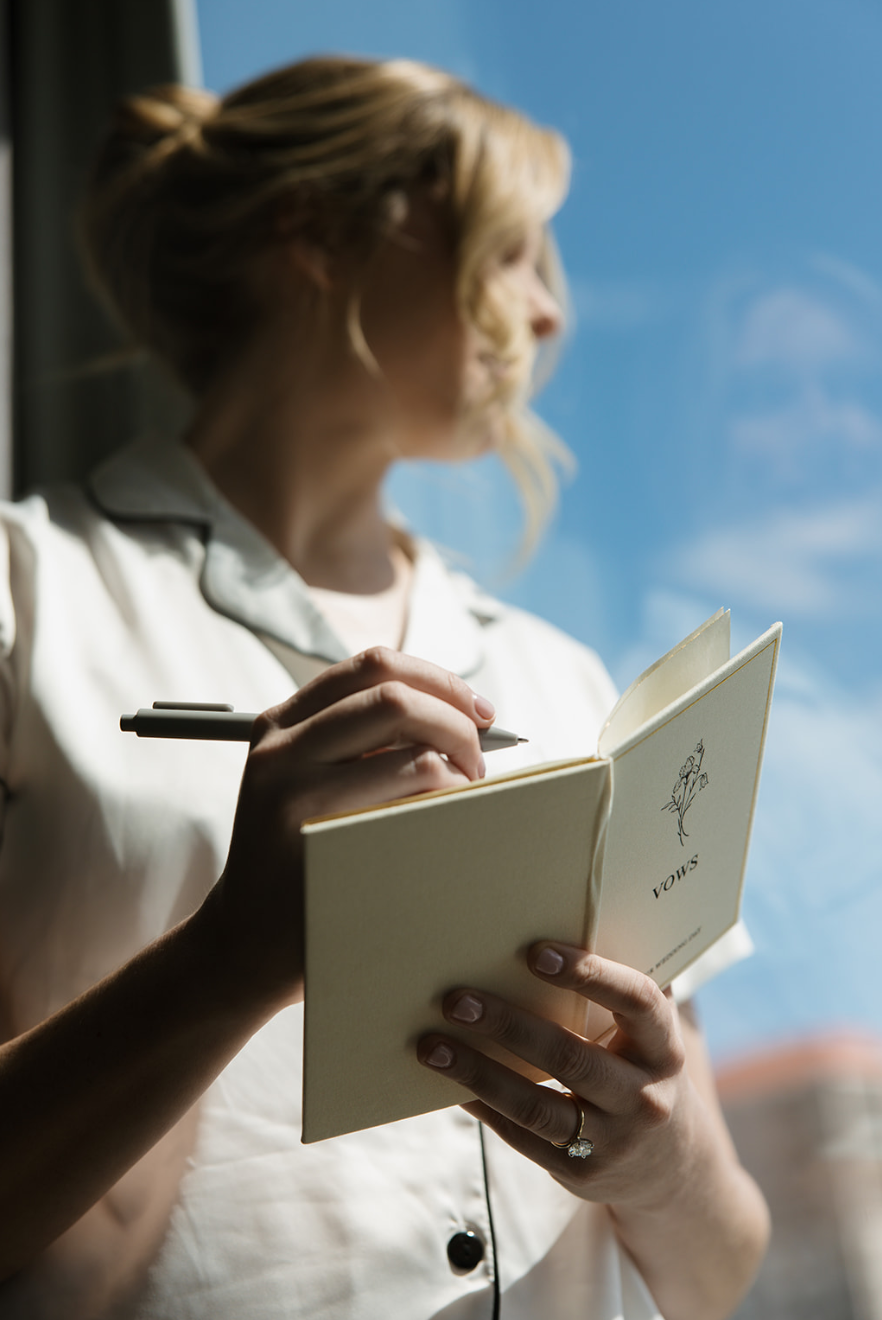 A woman with blonde hair wearing a white shirt is holding an open notebook and writing in it with a pen, standing near a window with blue sky and clouds visible outside.