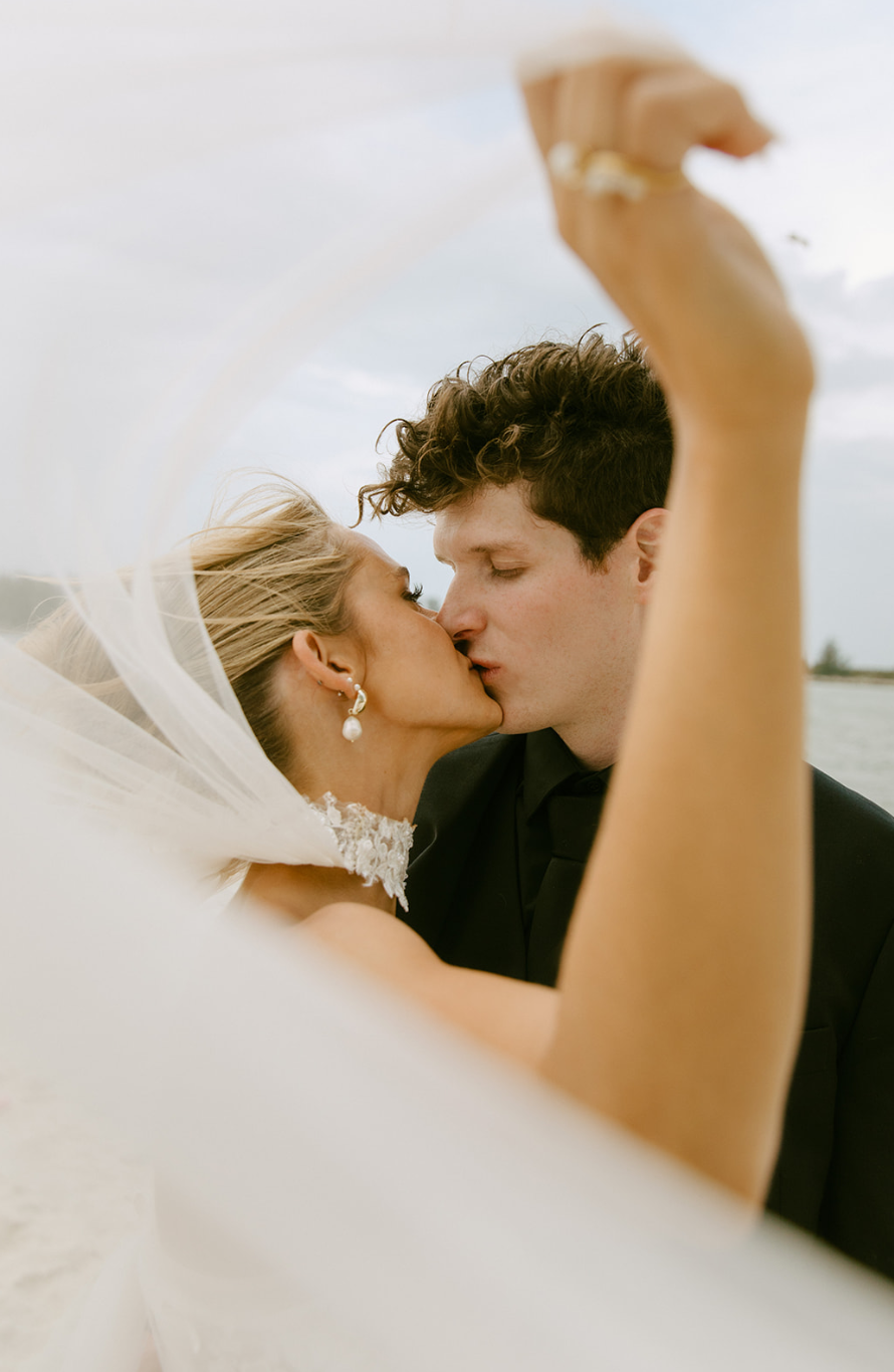 A bride and groom kissing outdoors, with the bride's veil flowing in the wind.
