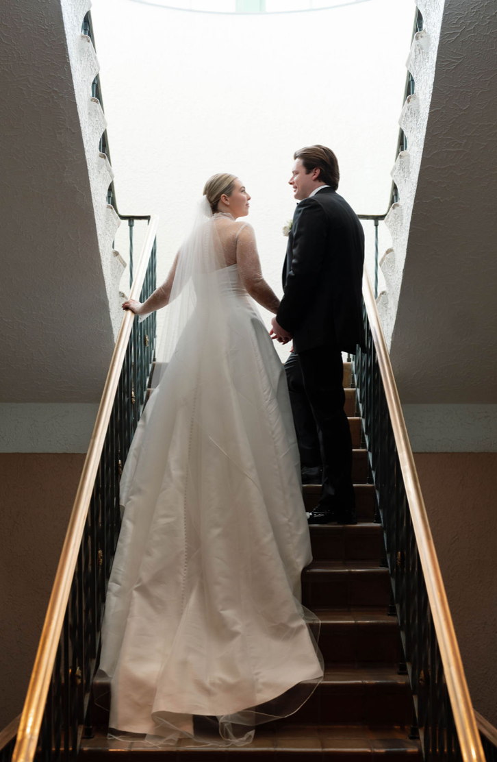 Bride and groom standing on staircase, holding hands, looking at each other, with the bride in a white wedding gown and veil, and the groom in a black suit, inside a building with a skylight ceiling.