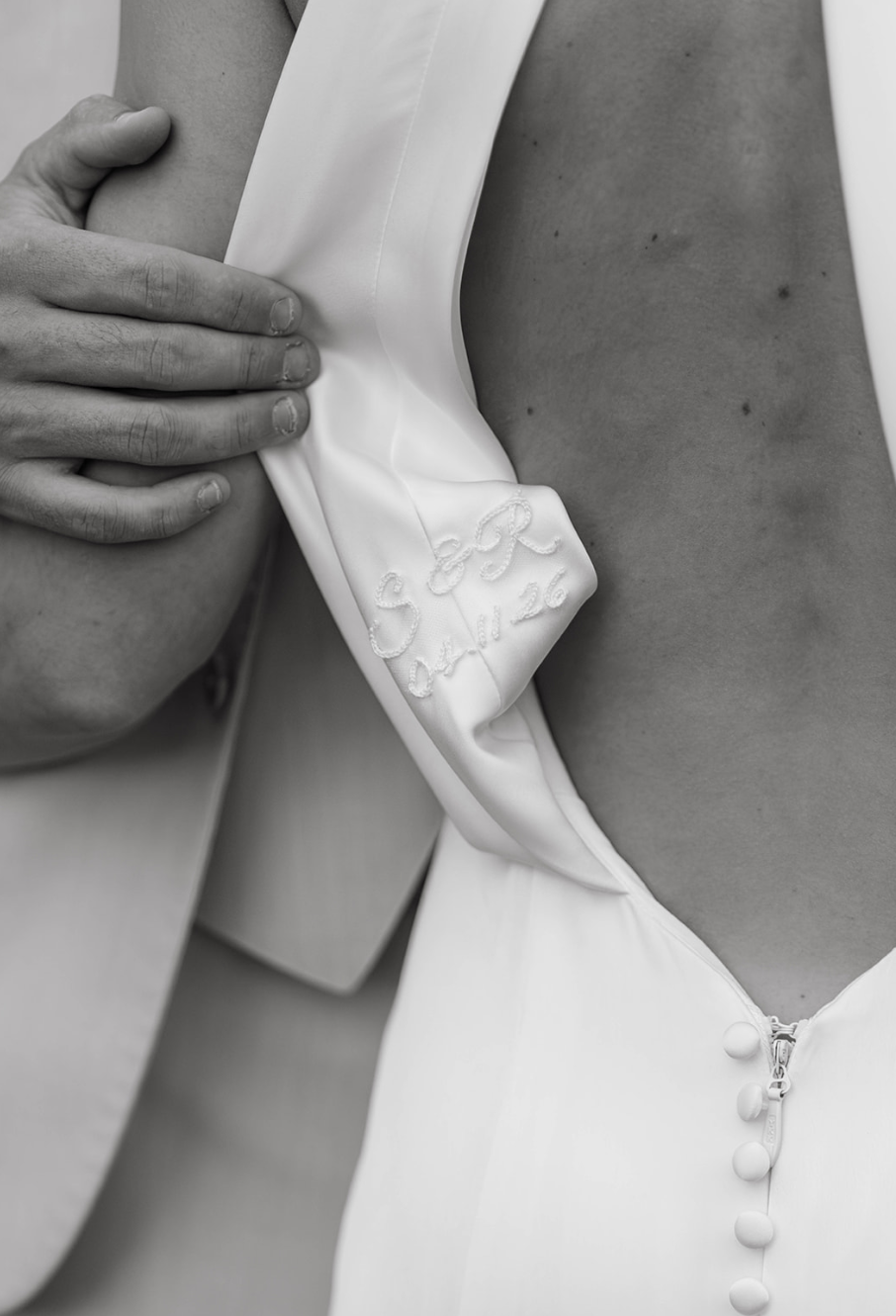 Close-up of a woman's chest revealing a white dress with a partially unzipped neckline. Someone's hand is pulling back the fabric, showing an embroidered message "G E R" with a date. The dress has buttons along the front.