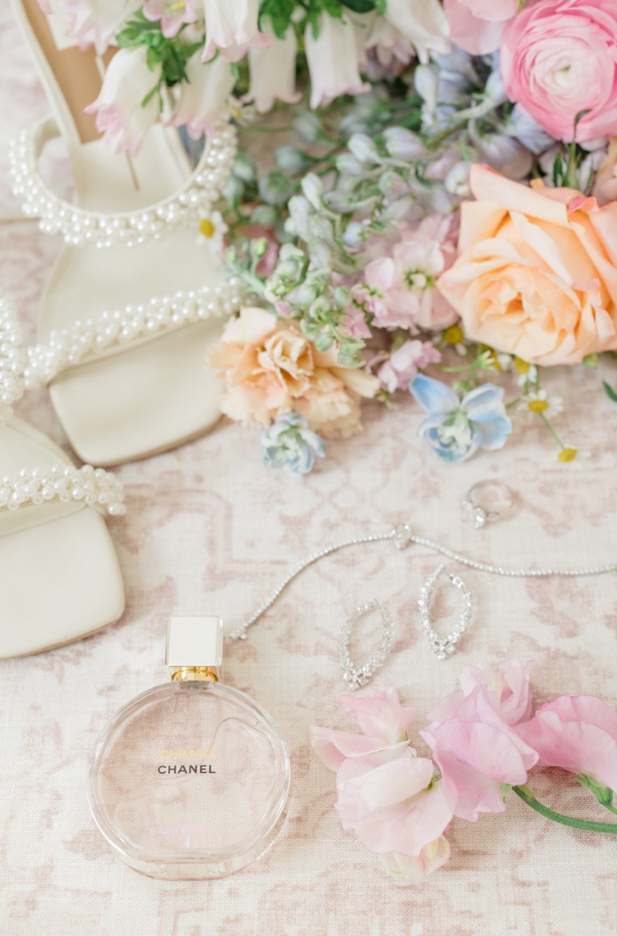 A collection of jewelry, a Chanel perfume bottle, and pastel pink and peach flowers arranged on a fabric surface.