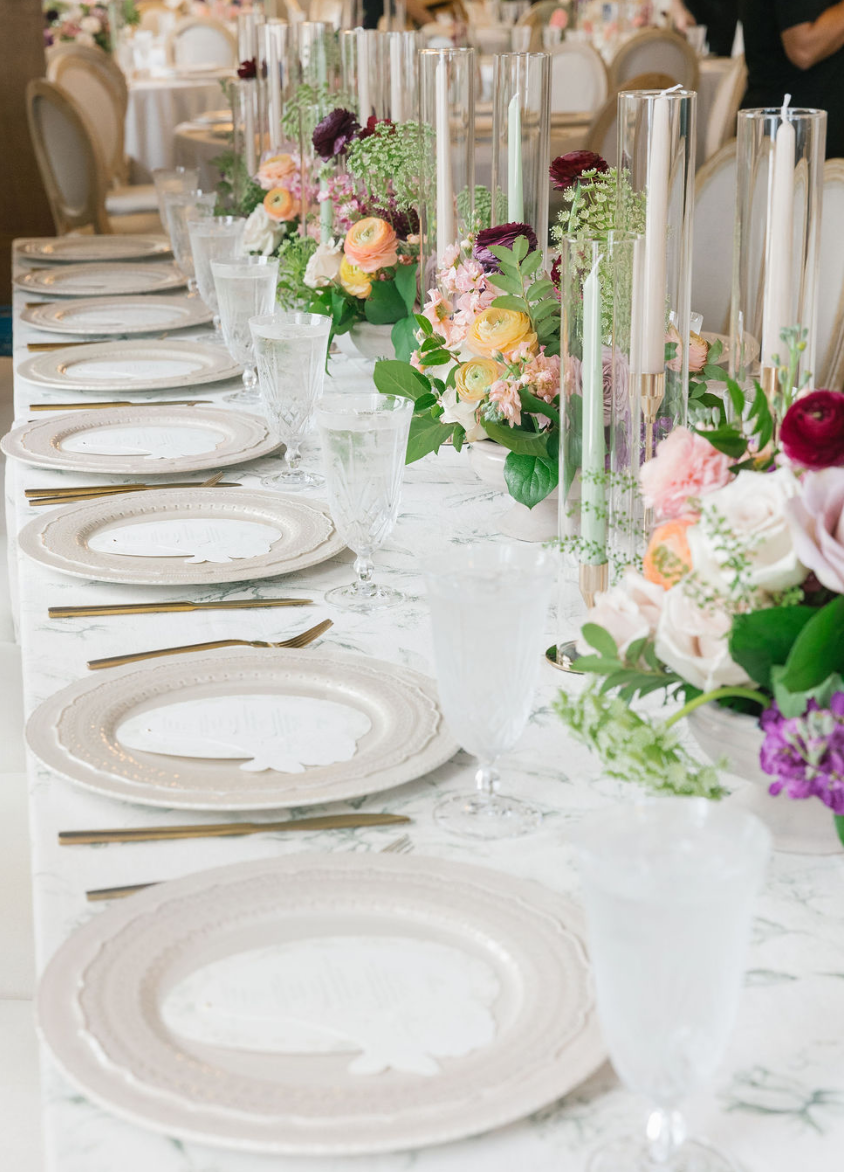 Elegant table setting with white plates, gold flatware, clear glassware, and floral centerpieces with pink, peach, and burgundy flowers in tall glass vases.