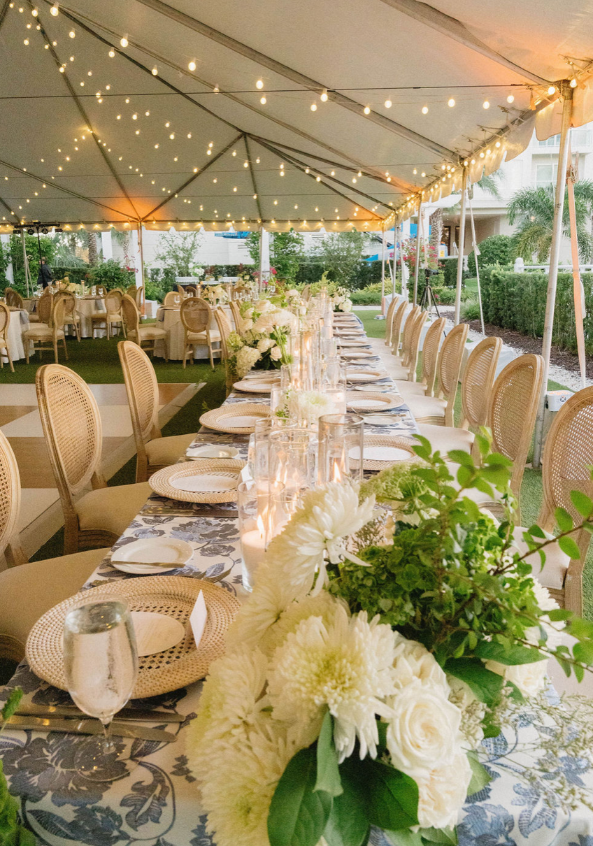 Long outdoor banquet table decorated with white flowers, candles, and glassware under a tent with string lights during daytime or early evening