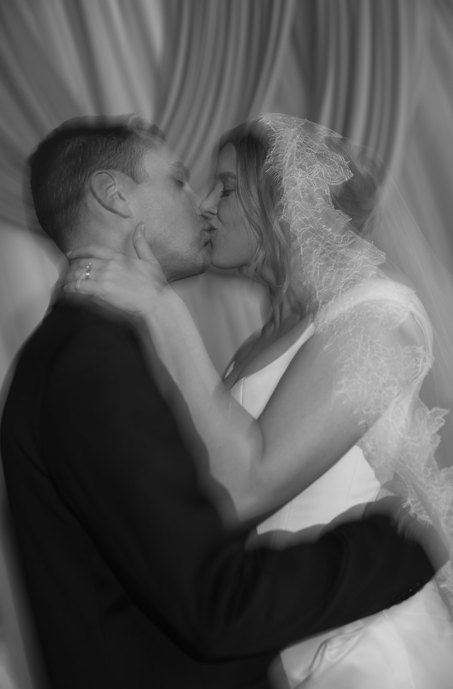 A black and white photo of a bride and groom kissing with a lace veil over the bride's head.
