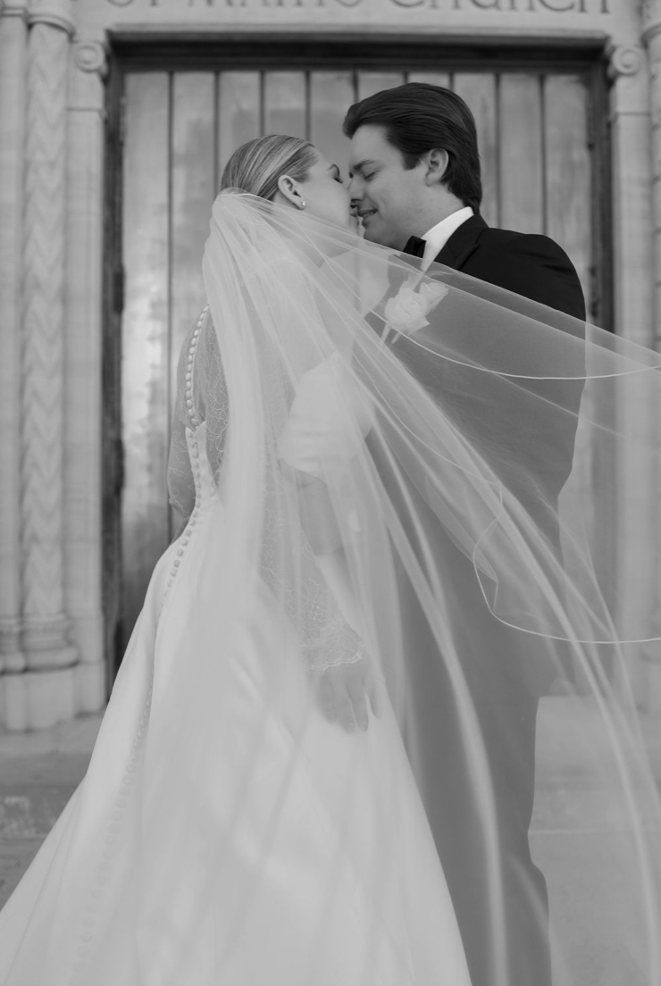 A black and white photo of a bride and groom embracing and about to kiss in front of a church entrance, with the bride wearing a veil and the groom in a tuxedo.