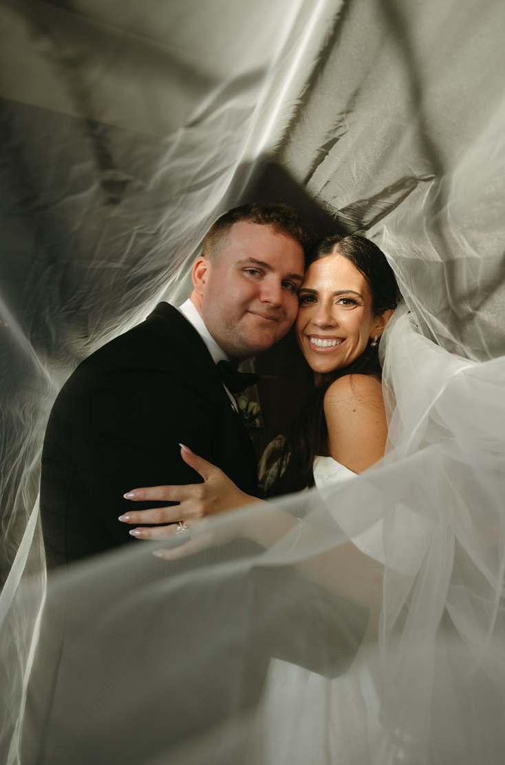 A newlywed couple smiling and embracing under a canopy of sheer white and gray fabric at their wedding.