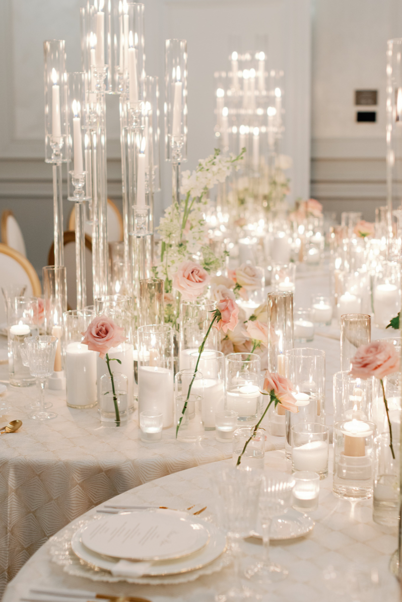 Elegant wedding table decorated with pink roses, numerous white candles in glass holders, and tall glass candelabras, set in a well-lit room with white walls.