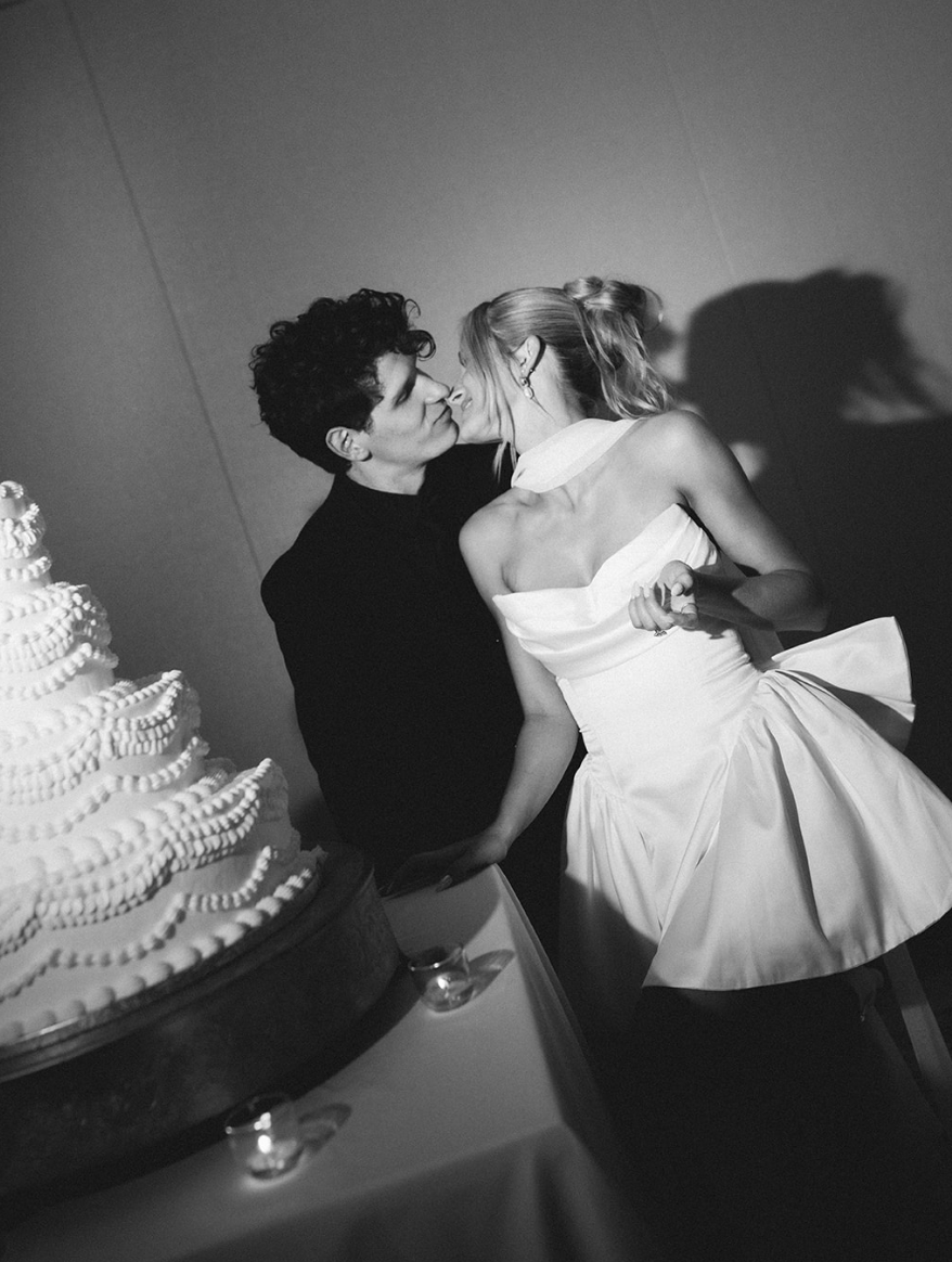 Black and white photo of a couple kissing at a wedding reception, with a tiered wedding cake decorated with white icing on the table in front of them.