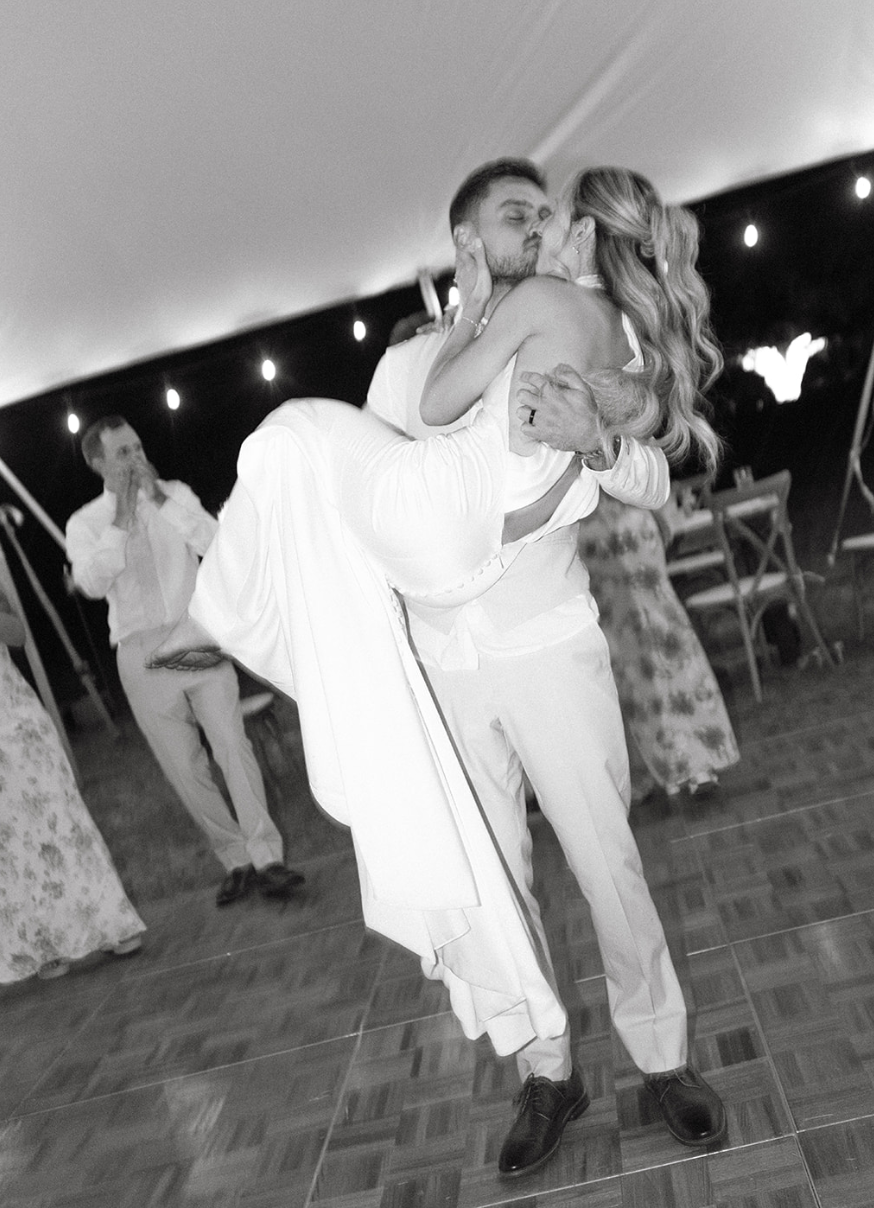 A man in a white suit is holding a woman in a white dress as they kiss at a wedding reception. The woman has long wavy hair, and the man is carrying her with one arm around her back and the other supporting her legs. In the background, for people are