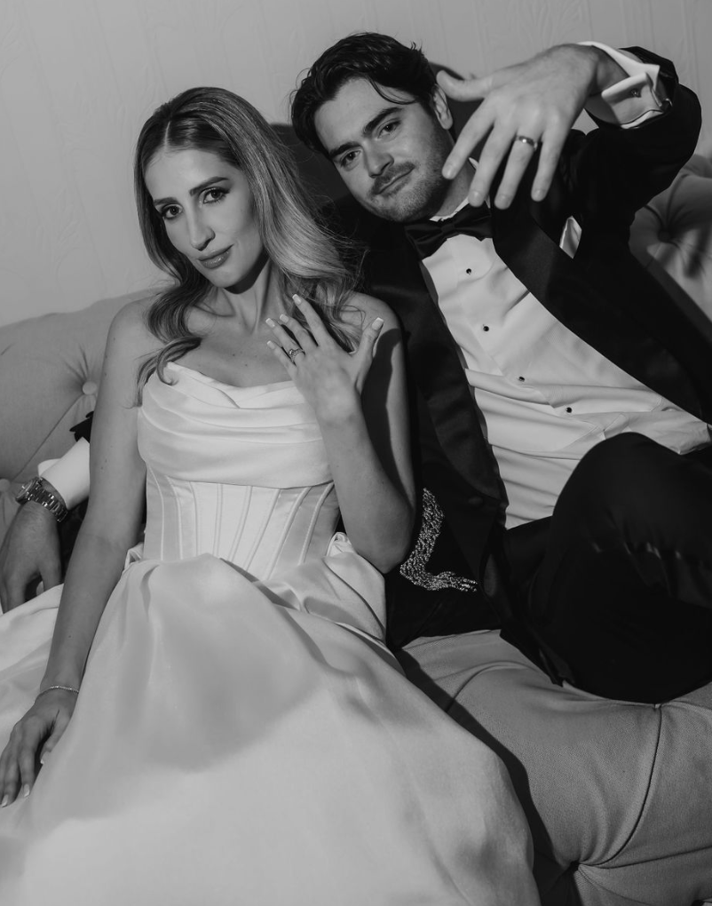 A black and white photo of a woman in an elegant dress and a man in a tuxedo sitting together on a couch. The woman displays her engagement ring, and the man is gesturing with his hand.