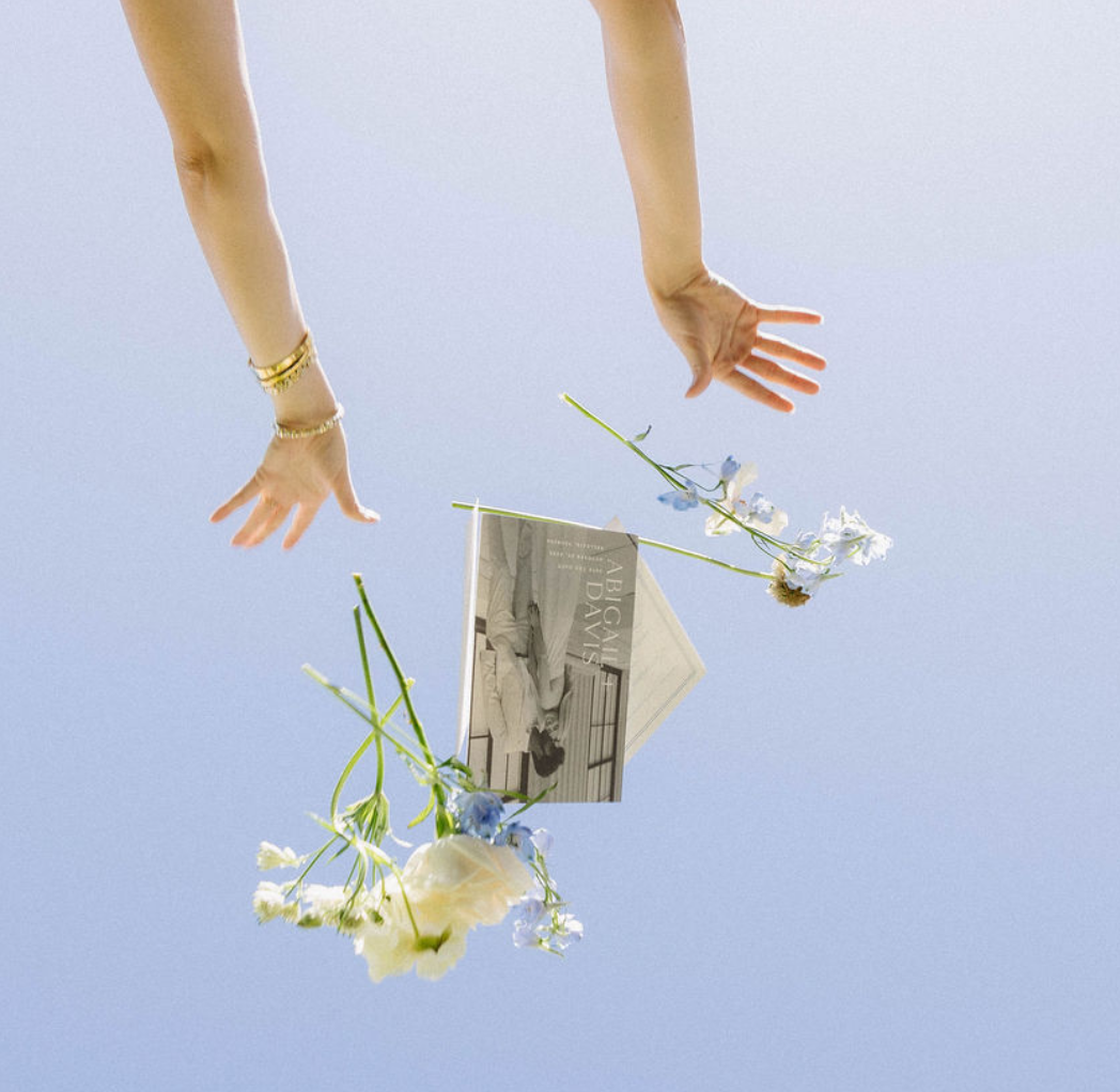 A person's hands with gold bracelets reaching toward floating white and blue flowers and a black-and-white magazine page against a light blue background.