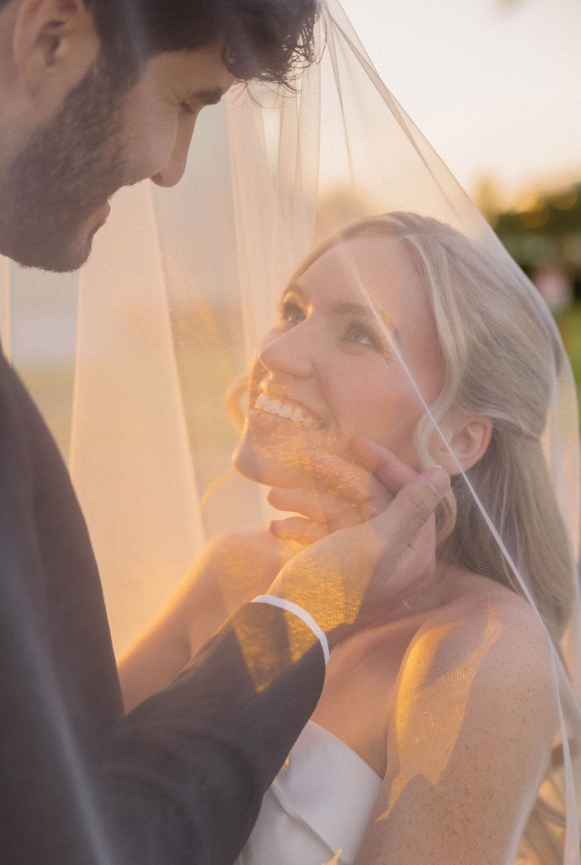 A bride and groom sharing an intimate moment under a wedding veil during sunset.