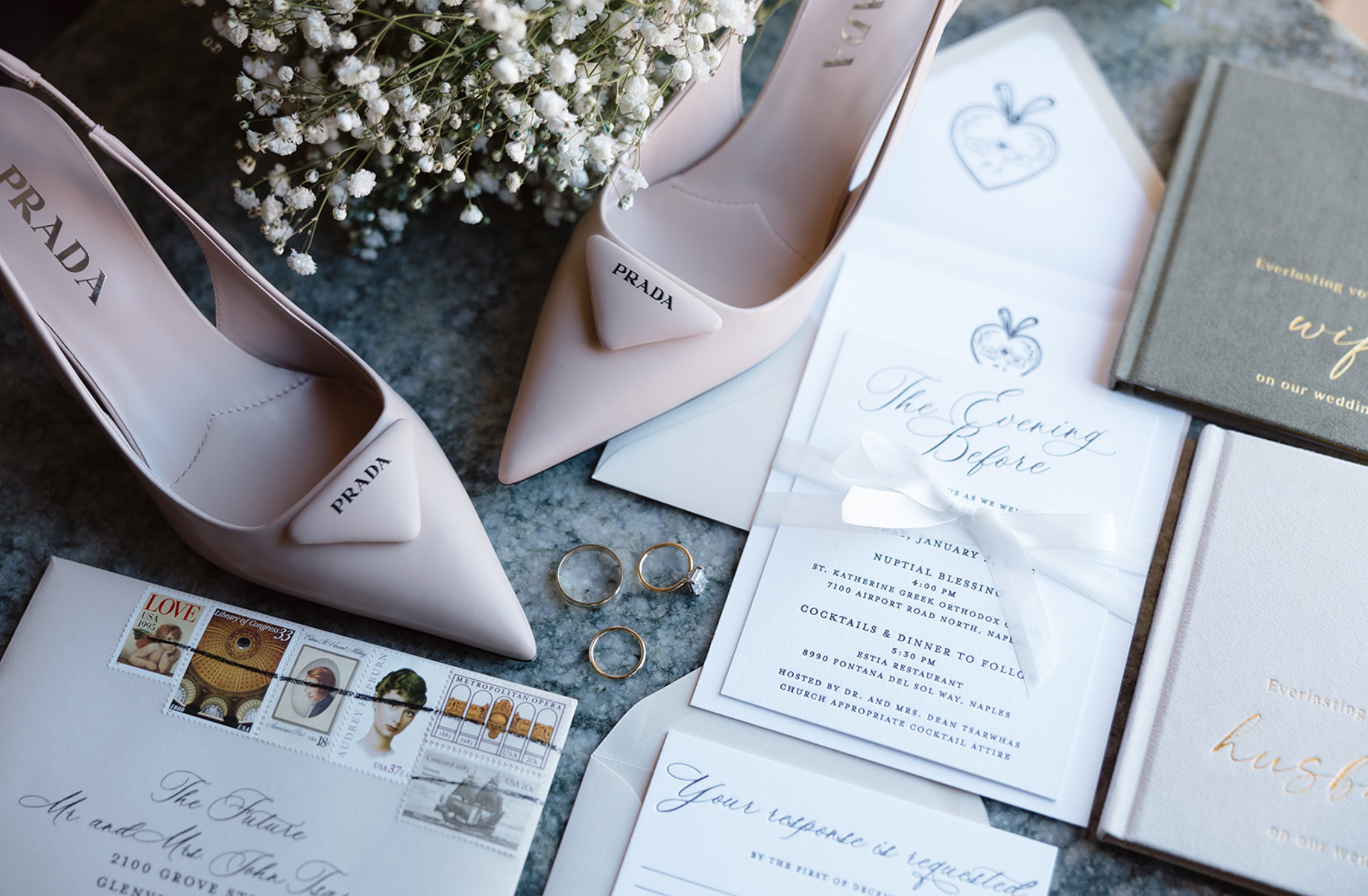 A pair of light pink Prada high-heeled shoes, white baby's breath flowers, wedding invitation cards, a small gray book, a greeting card with a strawberry illustration, wedding stamps, a gold wedding ring, and other wedding details arranged on a dark 