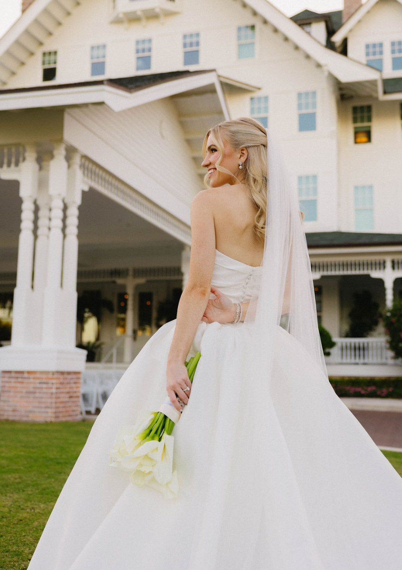 A smiling bride wearing a strapless white wedding gown and veil holding a bouquet of white calla lilies in front of a large white house with a wraparound porch.