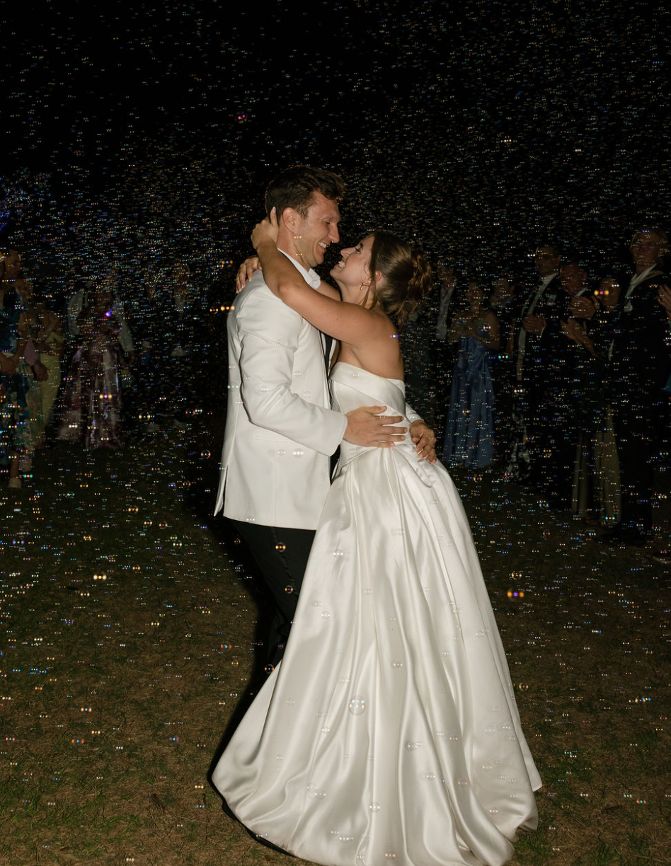 A bride and groom dancing at their wedding reception, surrounded by guests, at night with a starry sky background.
