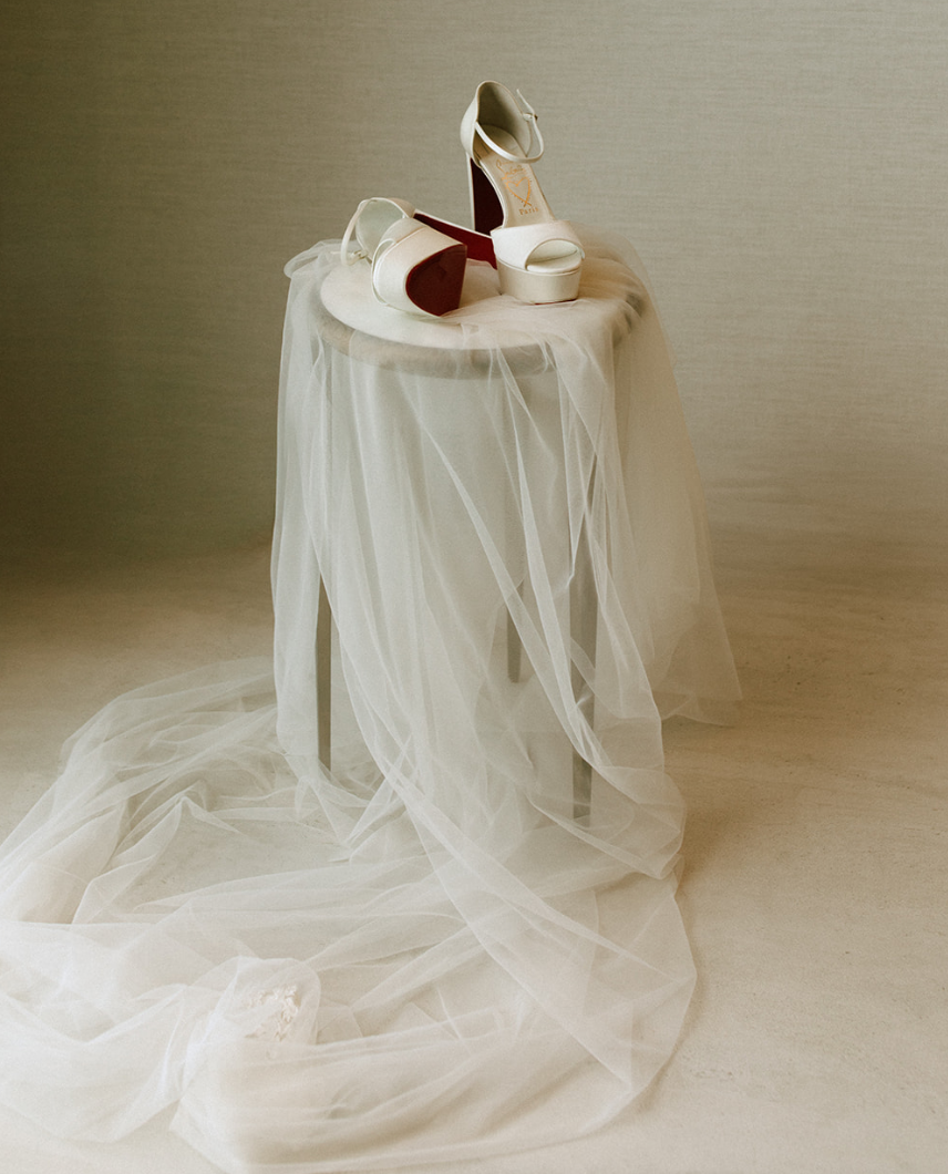 White high-heeled shoes with ankle straps placed on a small table draped with white tulle fabric, against a neutral background.