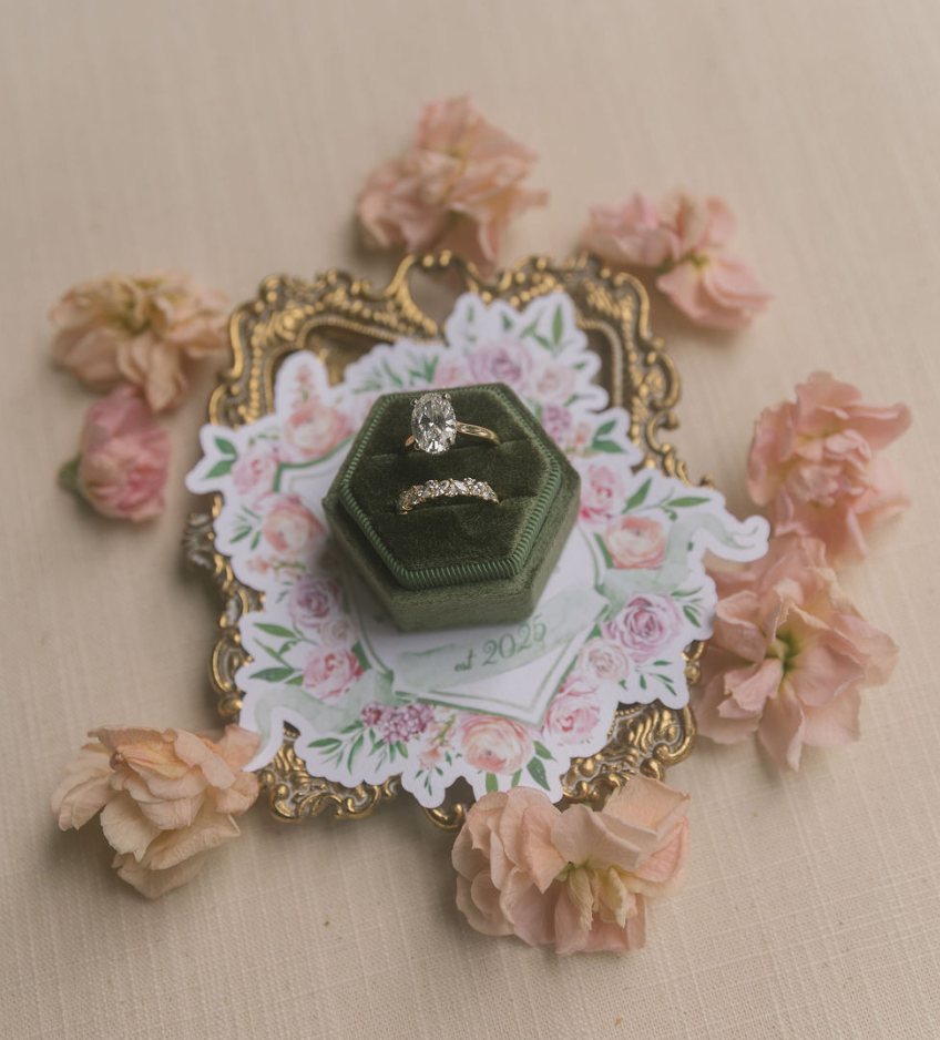 A gold ring with a large oval diamond, a gold and diamond band, and a floral card with pink flowers and green leaves, adorned with pink flower petals around it.