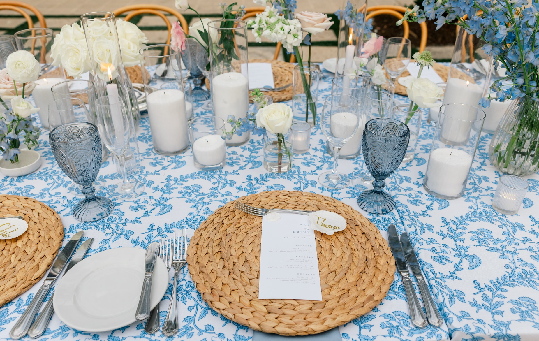 Decorated outdoor table with white and blue theme, featuring candles, floral centerpieces, woven placemats, and place cards for an event.