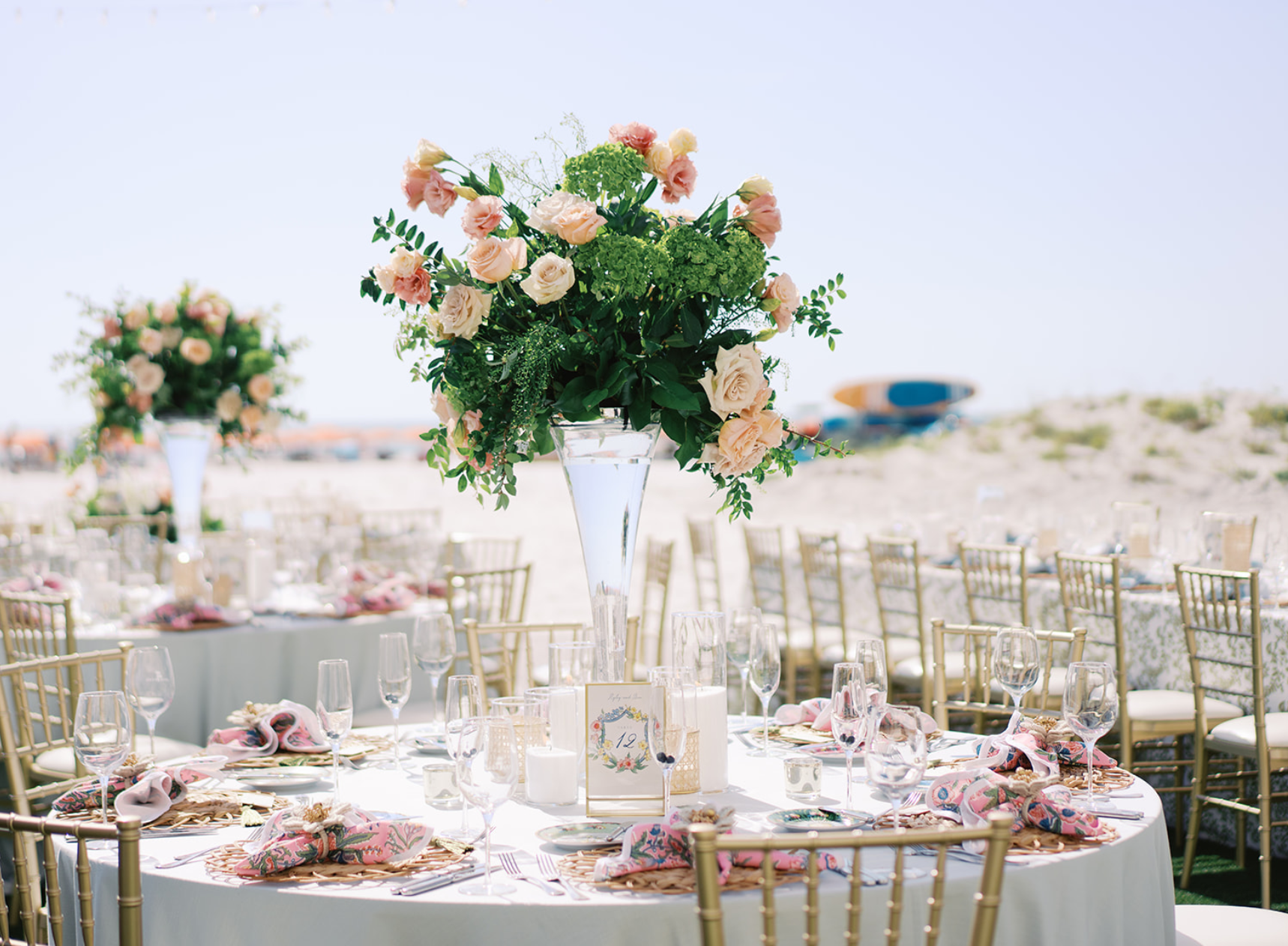 A large outdoor event setup with tables decorated with pink and white flowers, set on a sandy beach with chairs arranged around the tables, and a large floral centerpiece in a tall glass vase in the foreground.