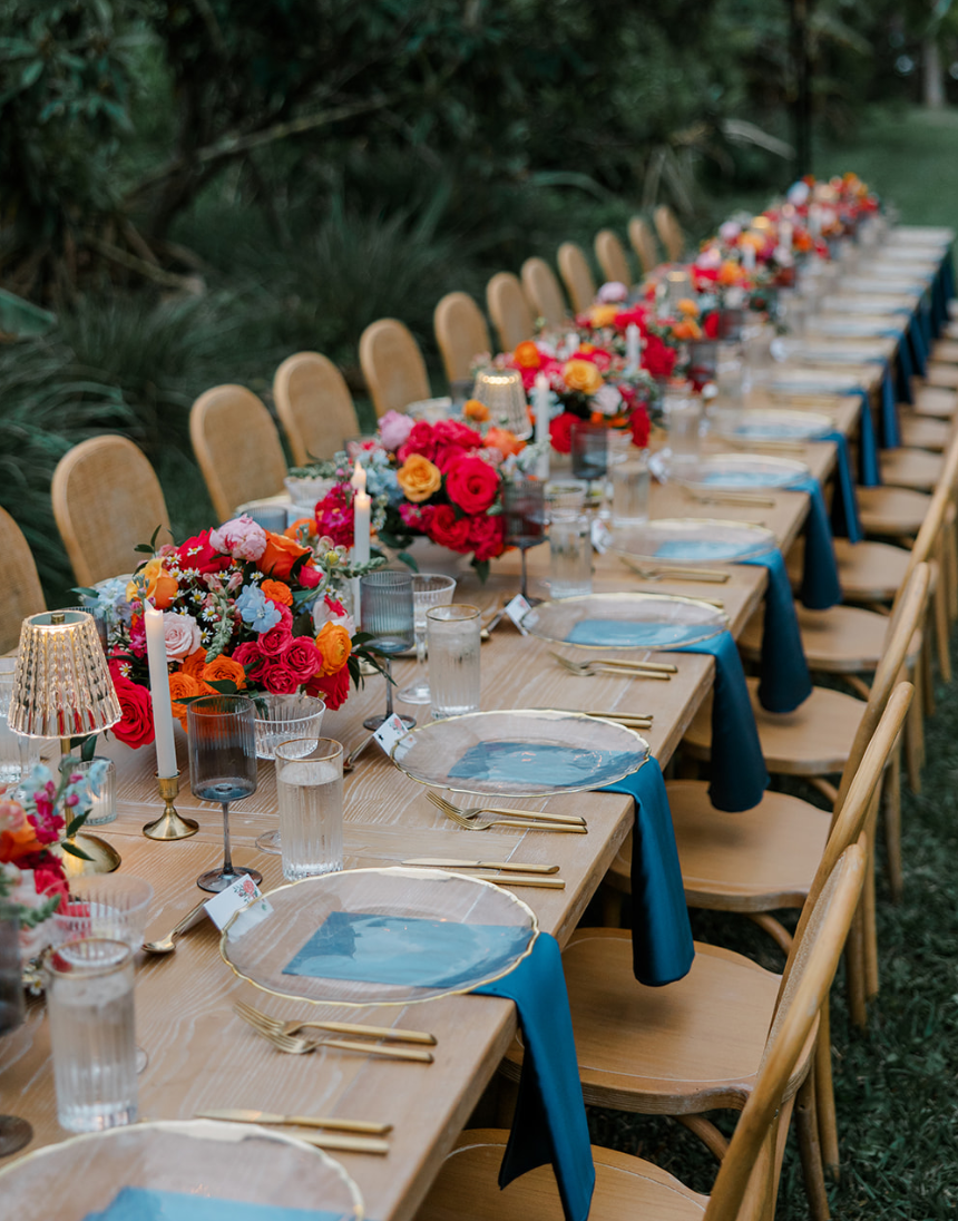 Long outdoor dining table set with colorful flower arrangements, candles, blue napkins, and elegant tableware in a garden setting.