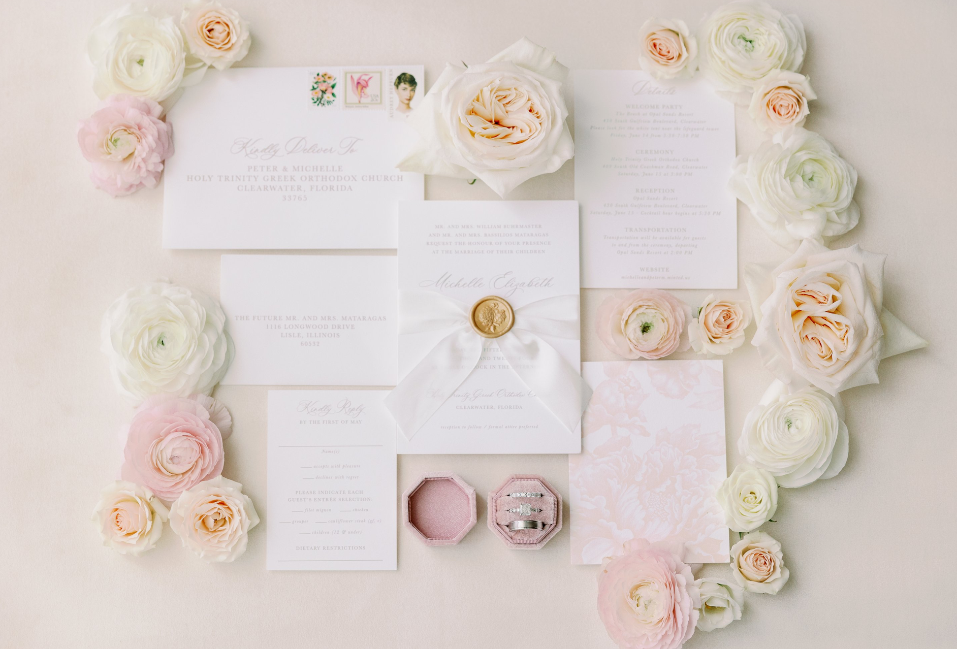 Wedding invitations and stationery surrounded by white and light pink roses, with two wedding rings in pink velvet ring boxes at the bottom center of the image.