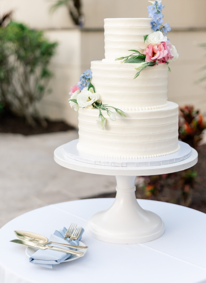 Three-tier white wedding cake decorated with pink, white, and blue flowers on a white cake stand. The cake is on a round table with a blue napkin, gold fork, and knife.