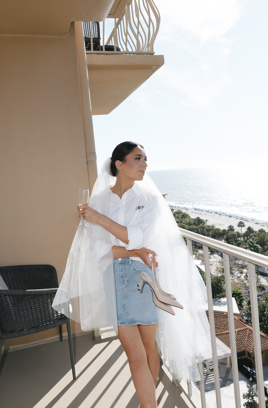 Woman in white shirt and denim skirt holding a pair of beige high heels and a glass of champagne on a balcony overlooking the ocean.