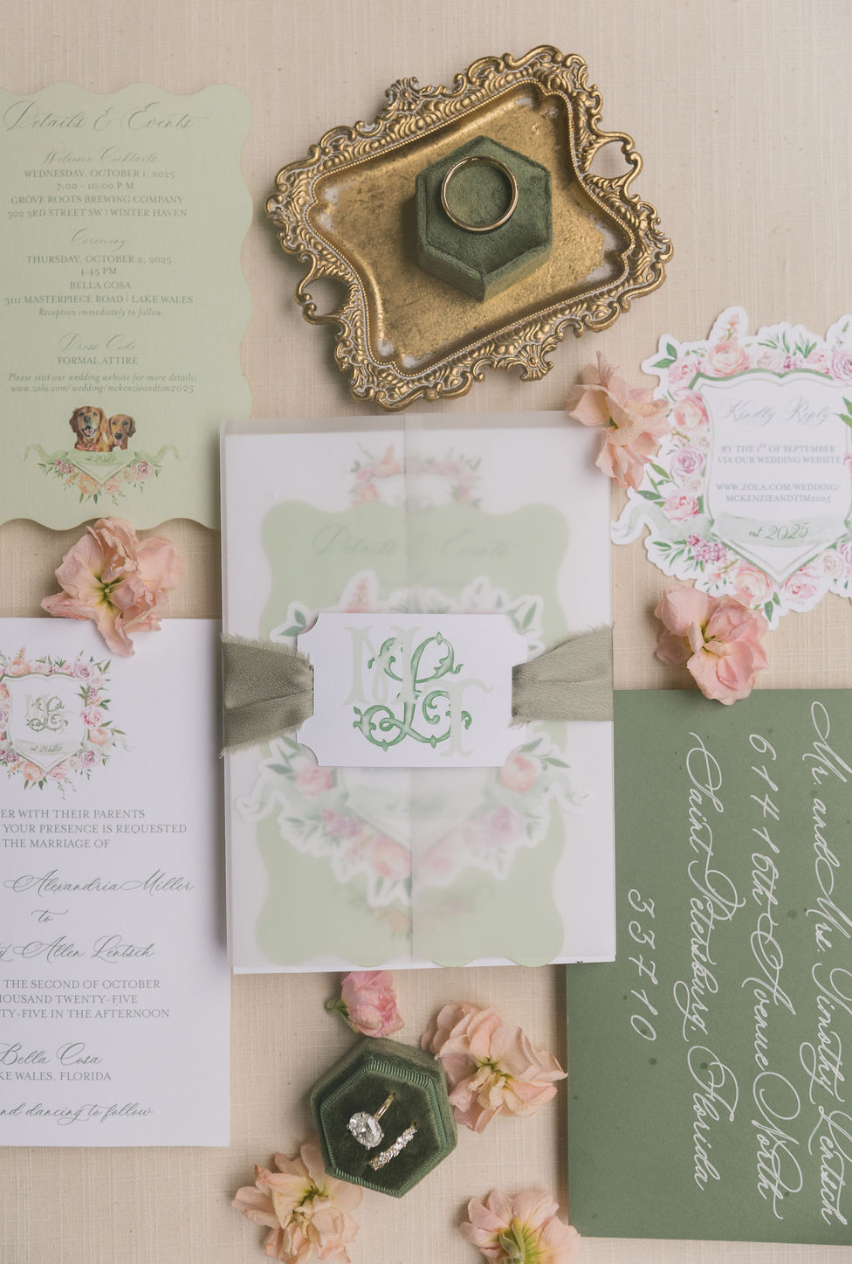 Wedding invitation suite with floral accents, pink flowers, and rings displayed on a decorative gold tray and in a green ring box.