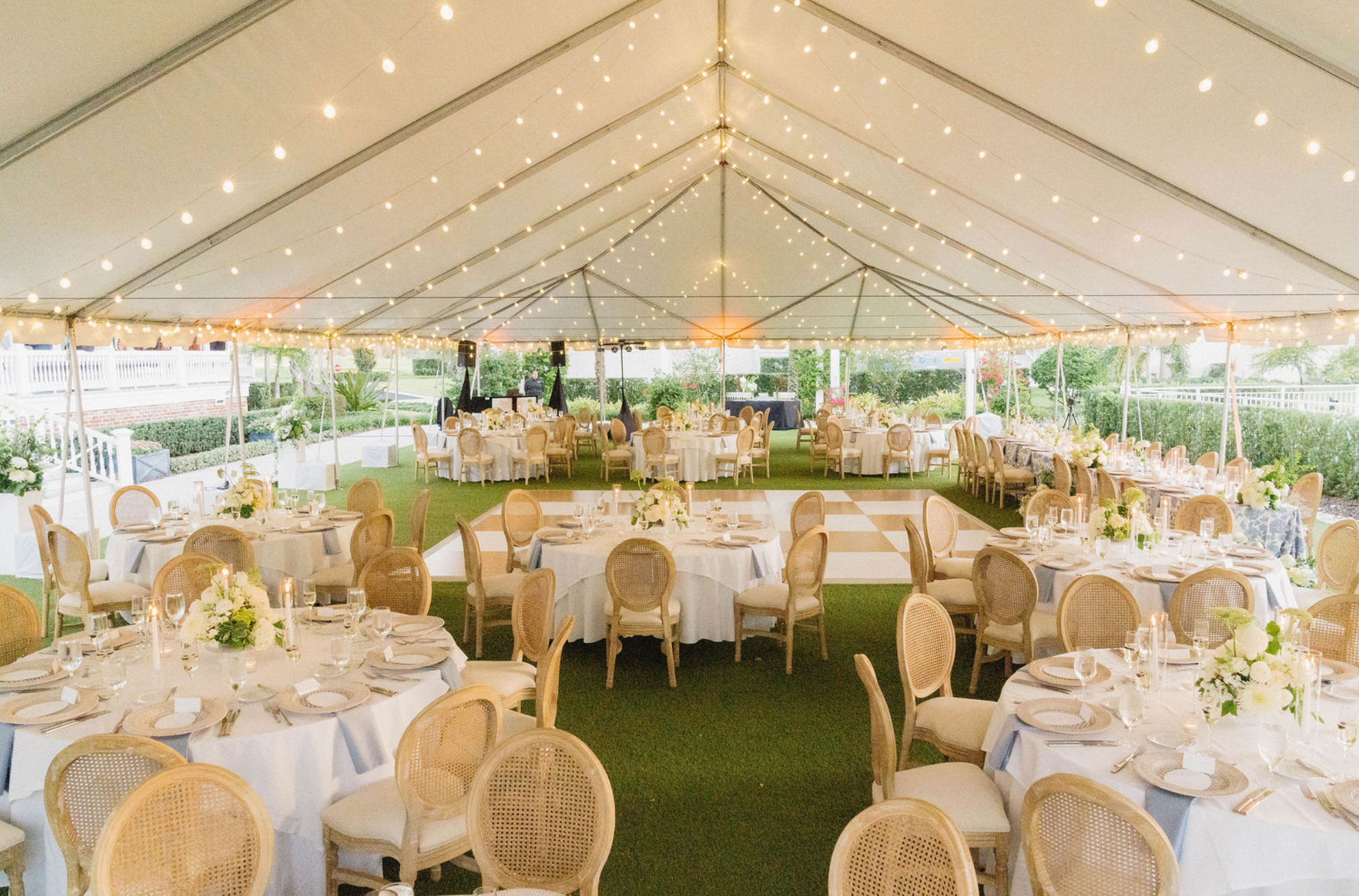 A large outdoor wedding reception tent decorated with string lights and floral centerpieces on round tables with white tablecloths, surrounded by beige chairs, on a grassy area with trees and bushes in the background.