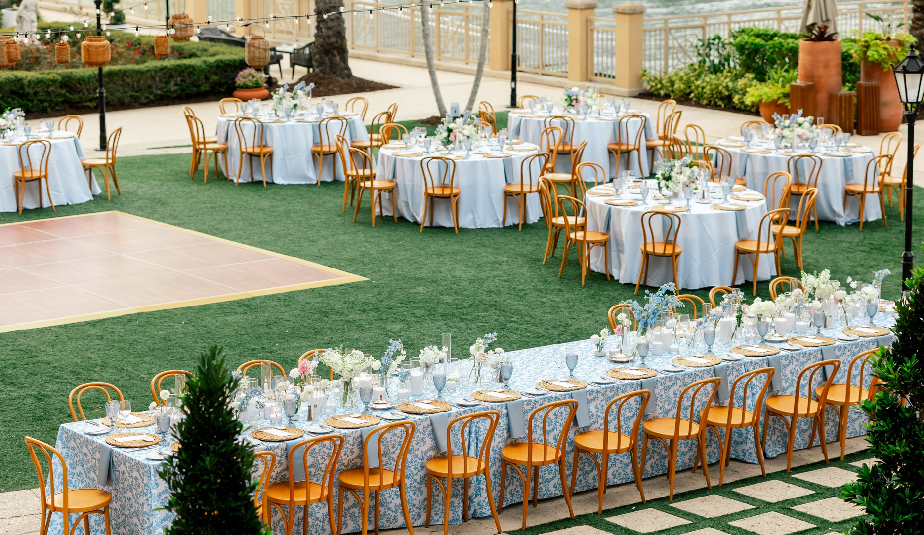 Outdoor wedding reception with round and long tables, decorated with white tablecloths, floral centerpieces, and place settings, in a garden setting with string lights, potted plants, and a dance floor.