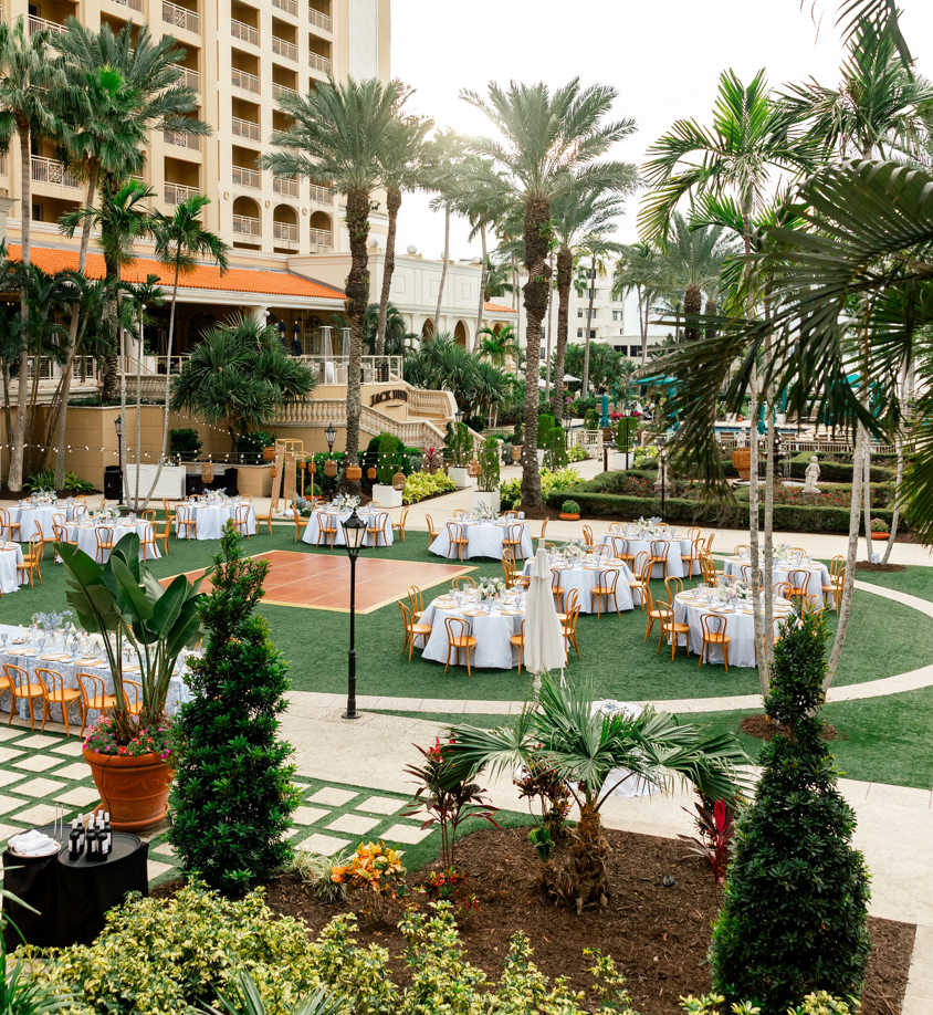 Outdoor event setup with round tables covered in white tablecloths, surrounded by wooden chairs, set on a grassy area with palm trees and lush plants, in front of a large hotel building.