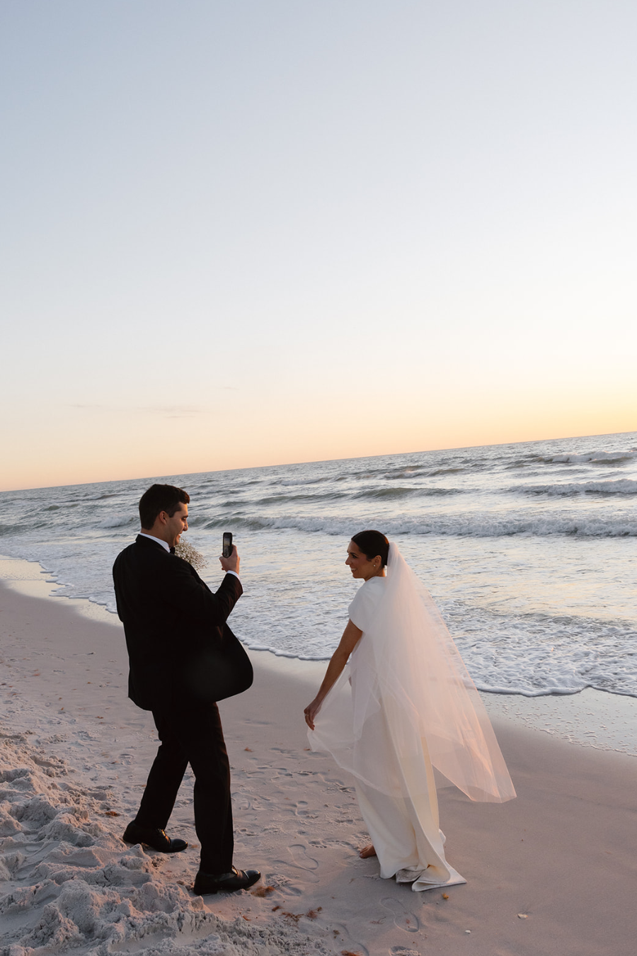 A bride in a wedding dress with a veil and a groom in a suit taking a photo together on a beach at sunset.
