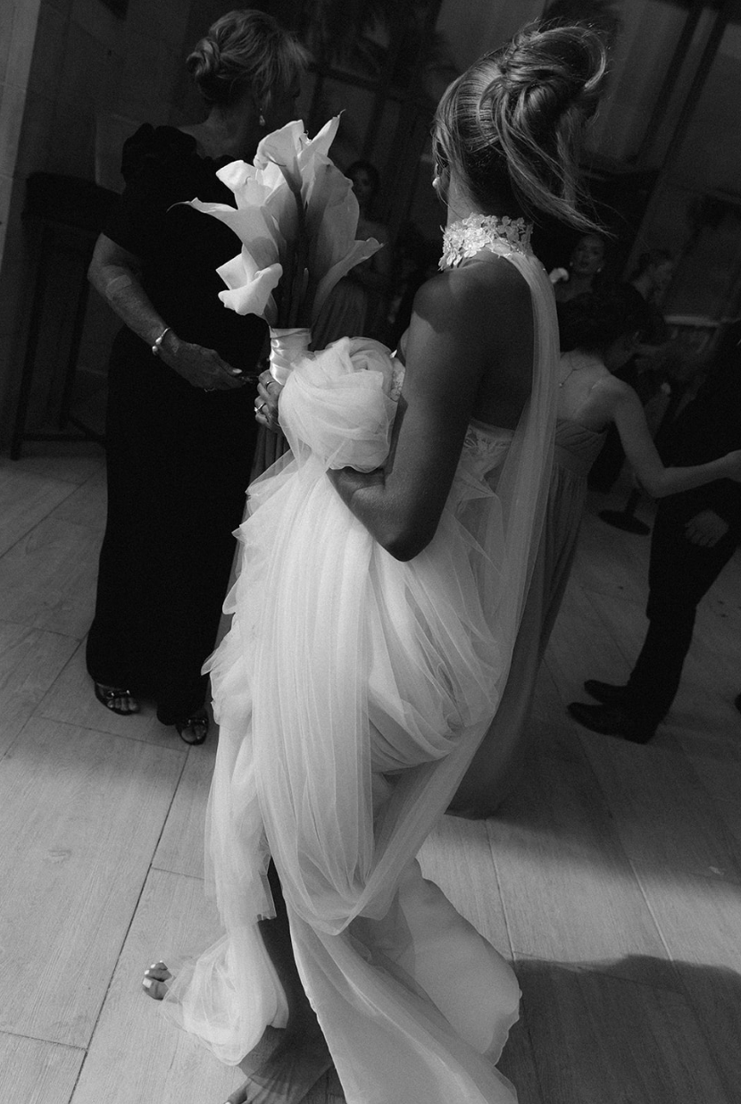 A bride holding a bouquet of calla lilies at a wedding reception, with guests in the background.