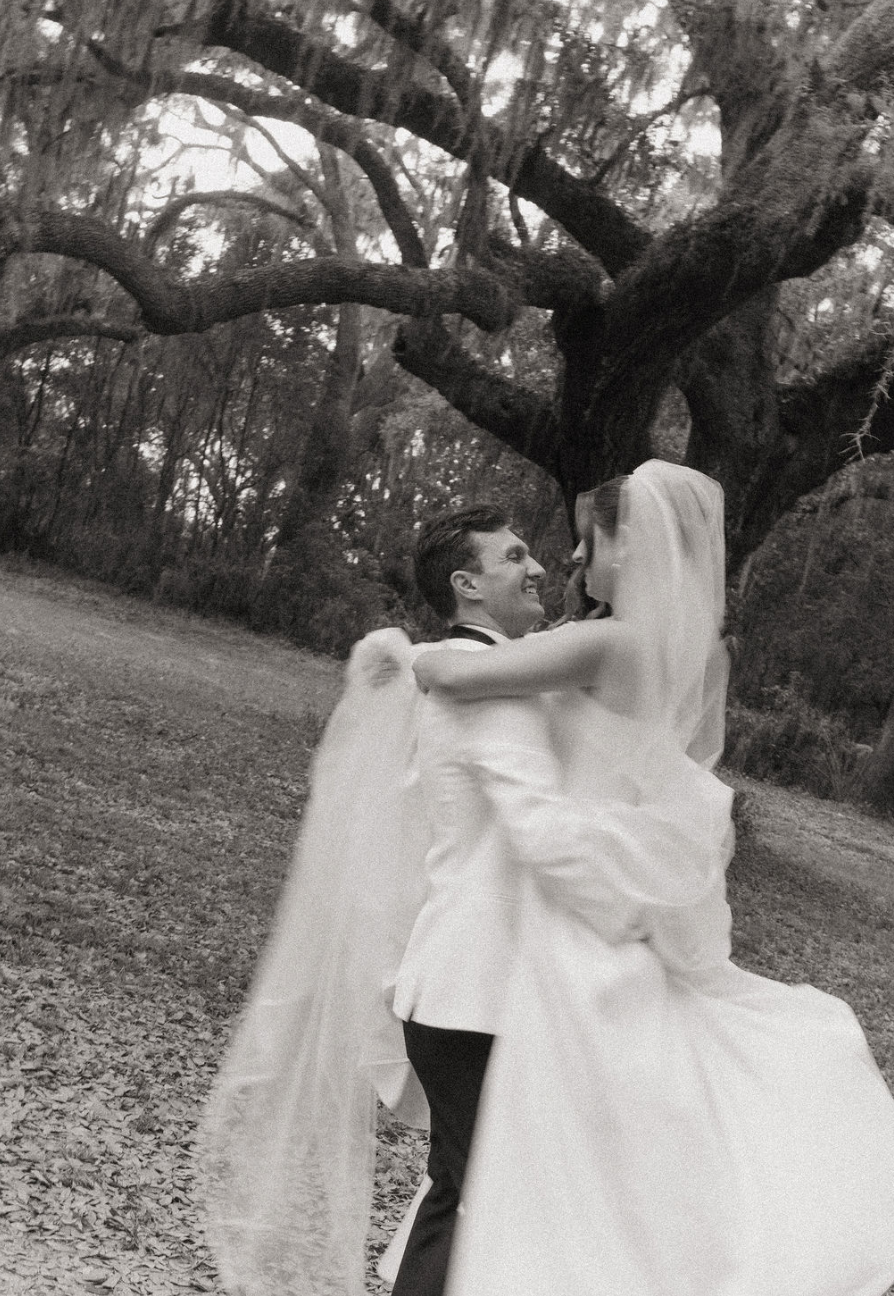 Black and white photo of a bride and groom dancing outdoors under a large tree, with the groom holding the bride up and both smiling.