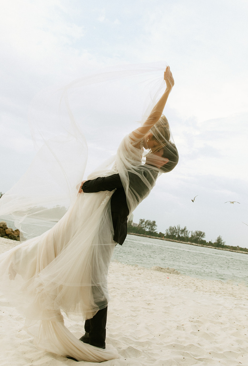 A couple kisses on a sandy beach, with the bride in a wedding gown holding a sheer veil and the groom in a black suit, with birds flying in the background.