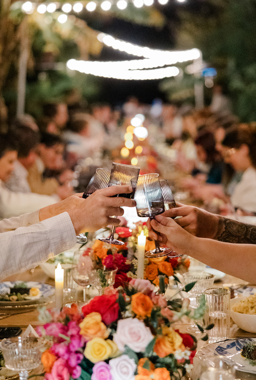 People at a dinner celebration raising glasses for a toast with a long table decorated with flowers and candles at night.