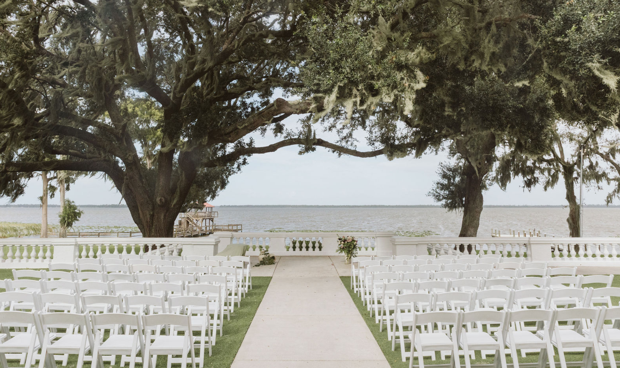 Outdoor wedding ceremony setup overlooking a lake, with white chairs arranged in rows on either side of a central aisle and large trees with moss hanging from the branches.