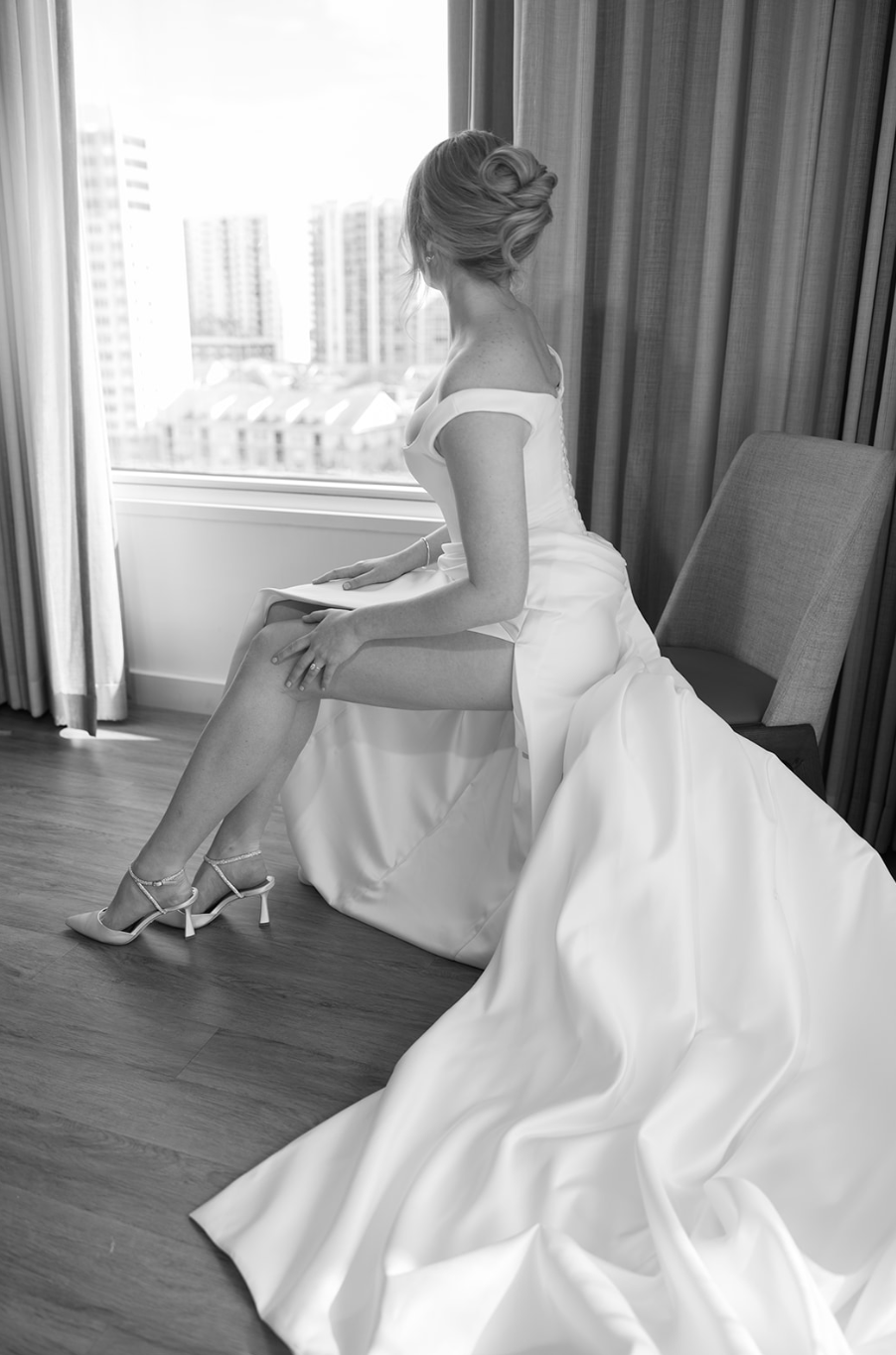 A bride sitting on a chair, wearing a wedding dress and high heels, looking out the window at city buildings.