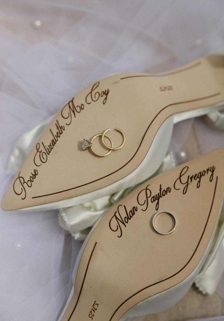 Close-up of a wedding ring and an engagement ring on a wedding seating chart with names Roe Elizabeth Mo Coy and Nalan Payton Gregory written in elegant script.